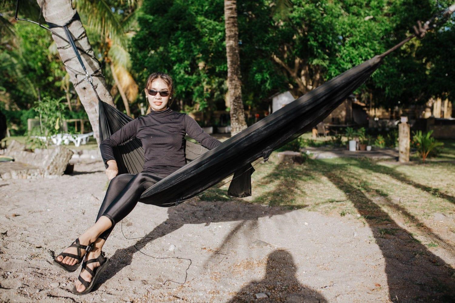 Woman in black clothes relaxing on a black hammock at a sunny tropical beach garden