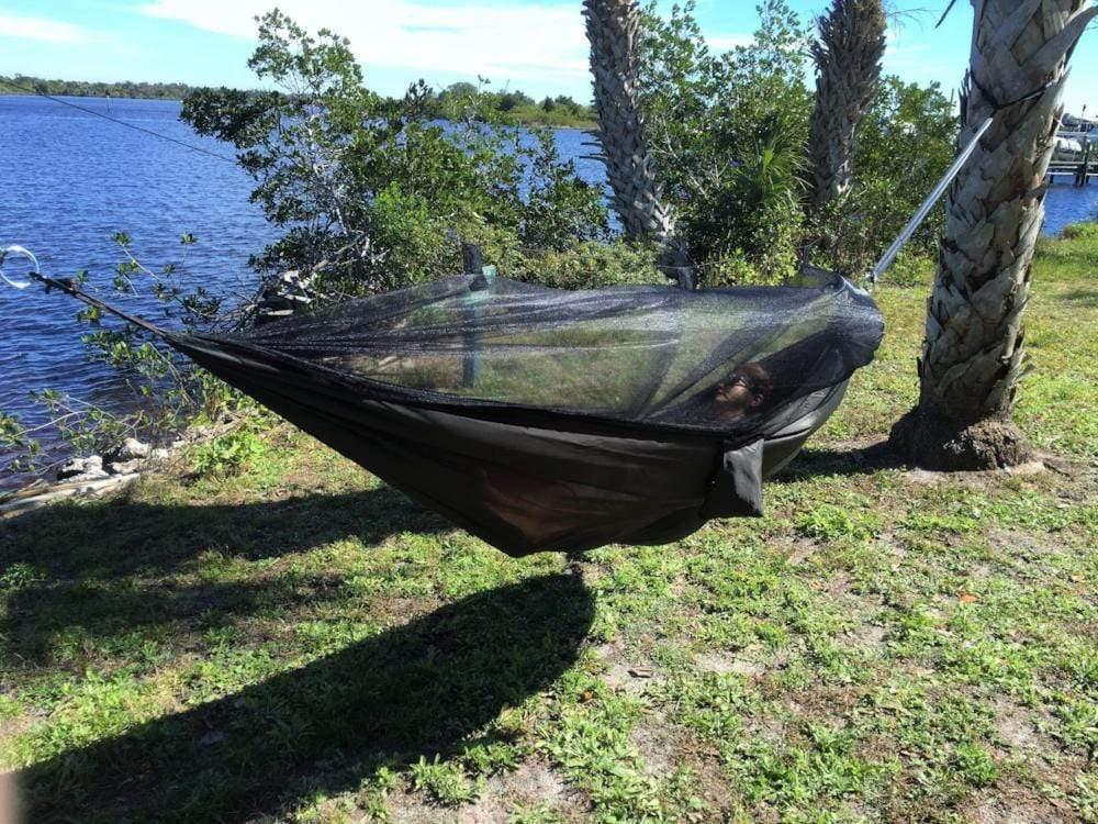 Dark green camping hammock with mosquito net by a lake, tied between palm trees outdoors