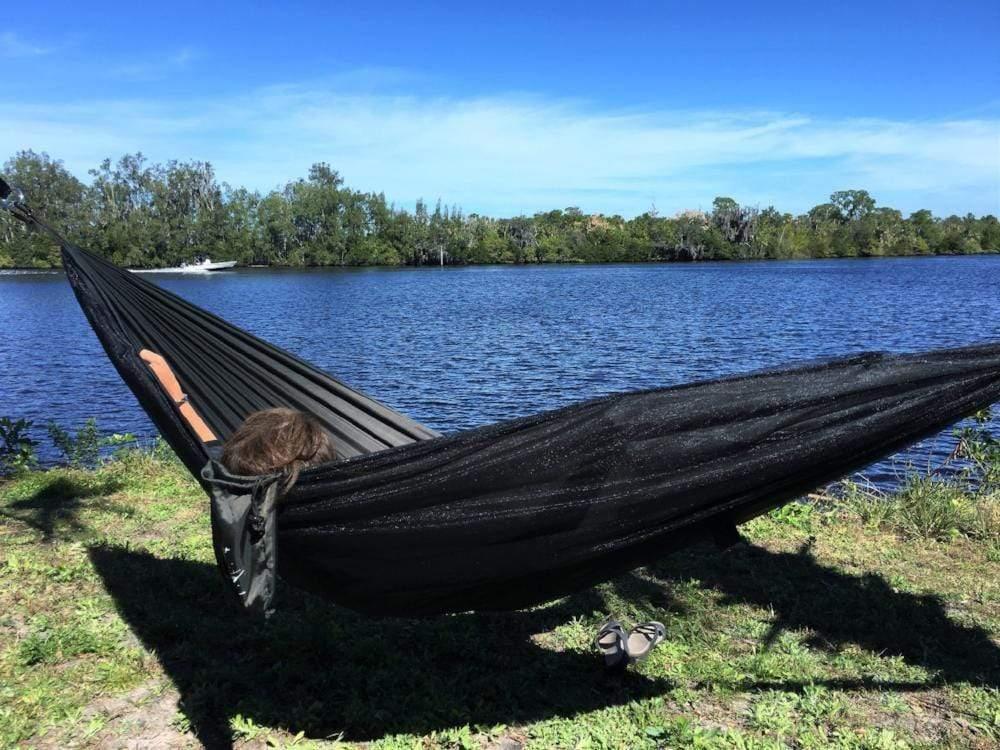 Dark green hammock with person relaxing by a river on a sunny day, outdoor nature scene