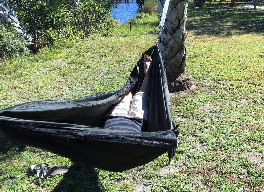 Person relaxing in dark green hammock tied to tree outdoors by grassy area
