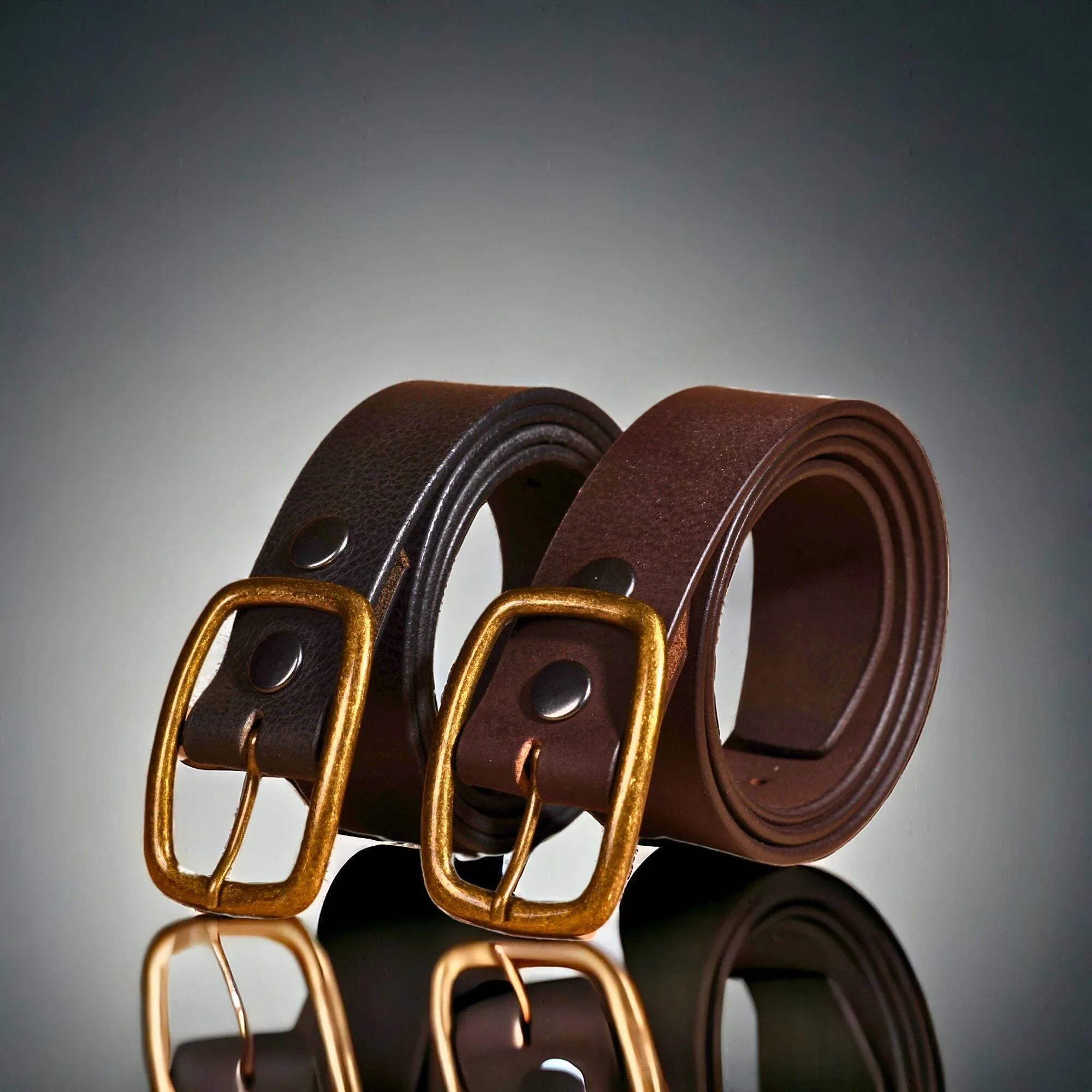 Two brown leather belts with brass buckles on a reflective surface
