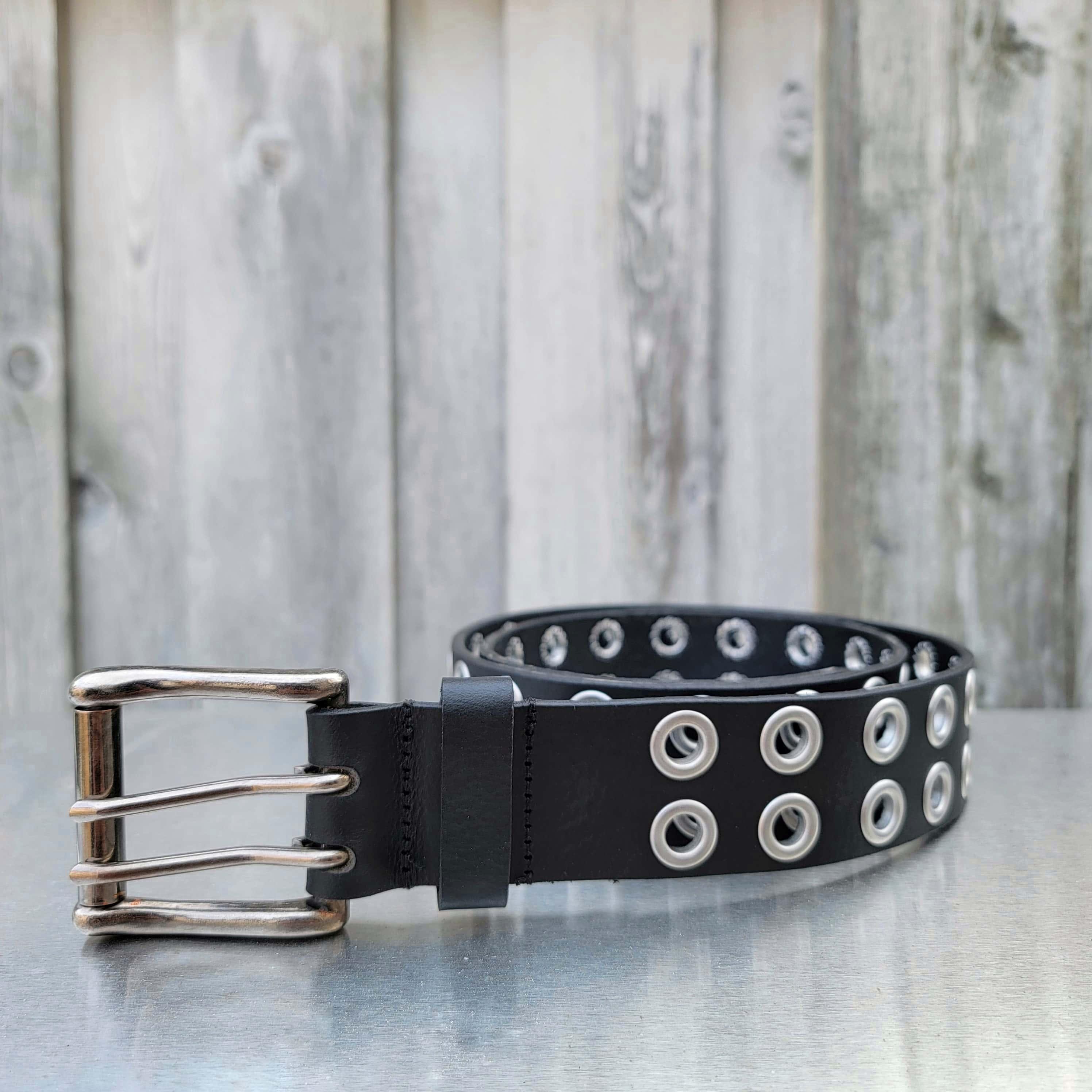 Black leather double grommet belt with silver buckle on metal surface, wood background