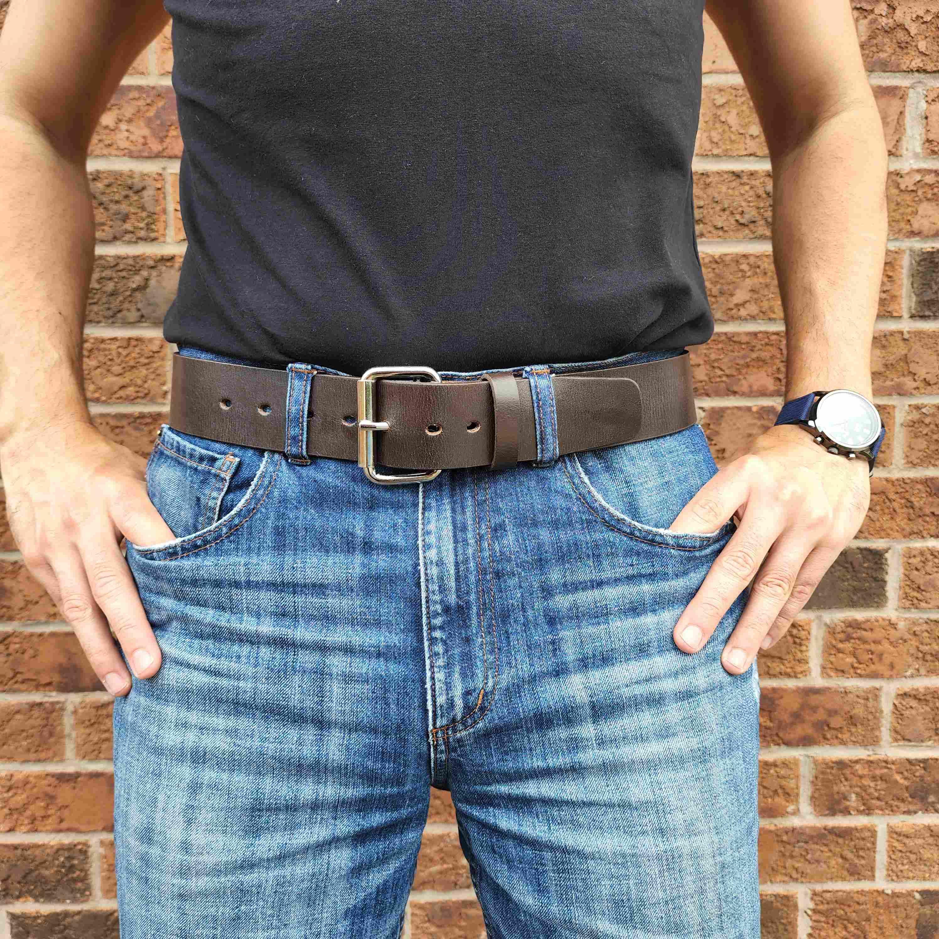 Man wearing black shirt, brown leather belt, blue jeans, and watch against brick wall