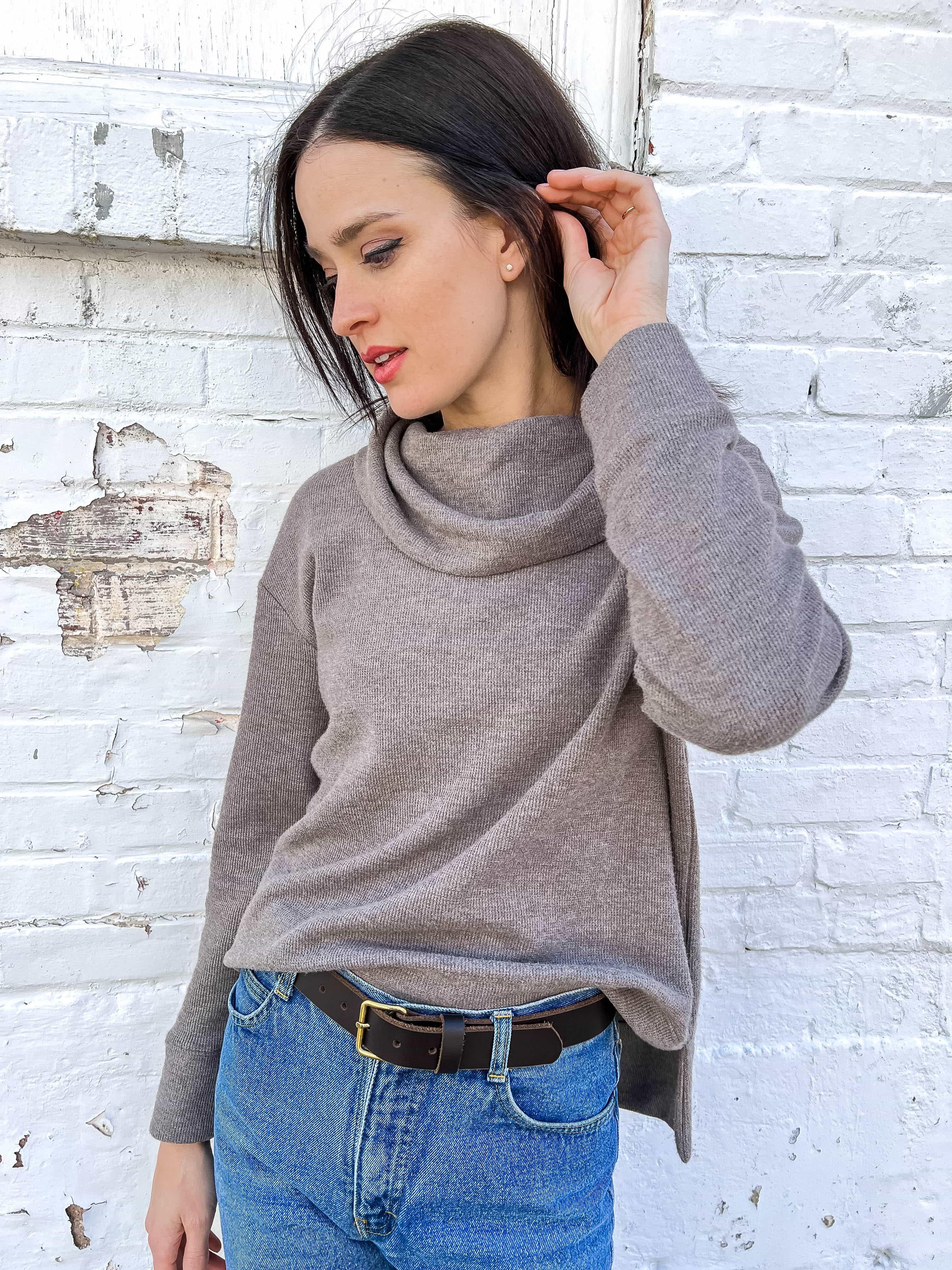 Woman in taupe cowl neck sweater and blue jeans posing against a white brick wall
