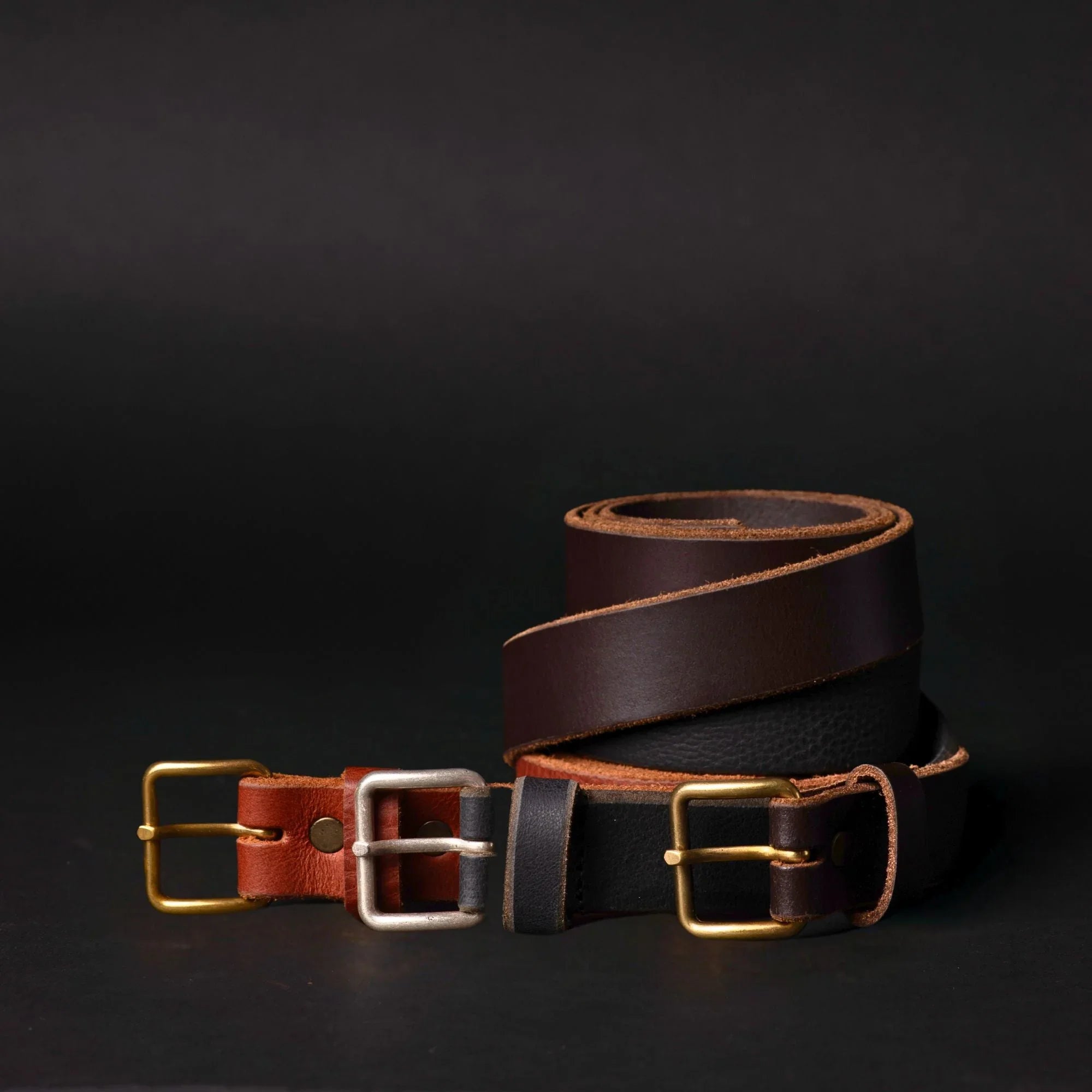 Rolled brown and black leather belts with metal buckles on black background