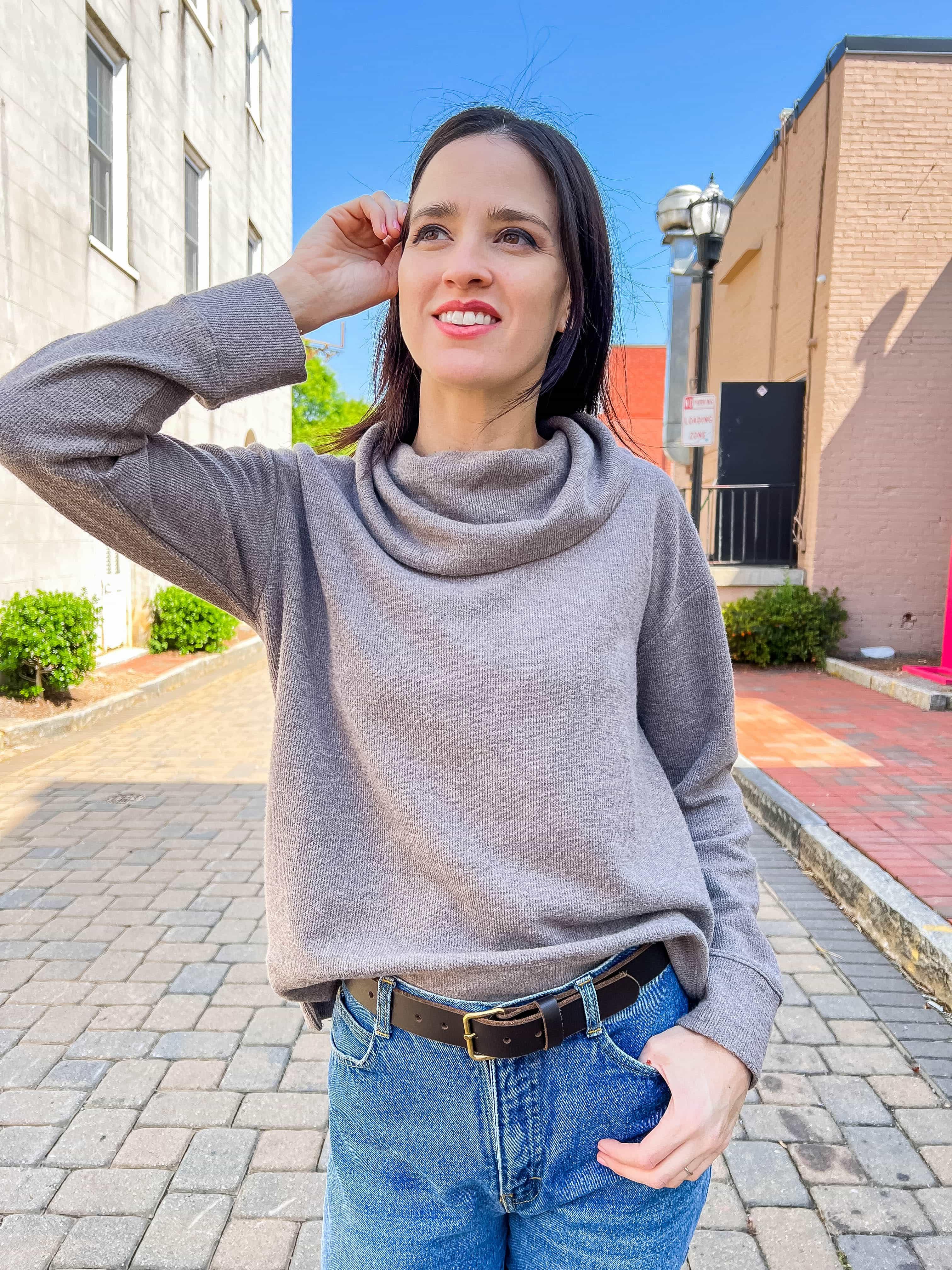 Woman in gray cowl neck sweater and jeans smiling outdoors on sunny city street