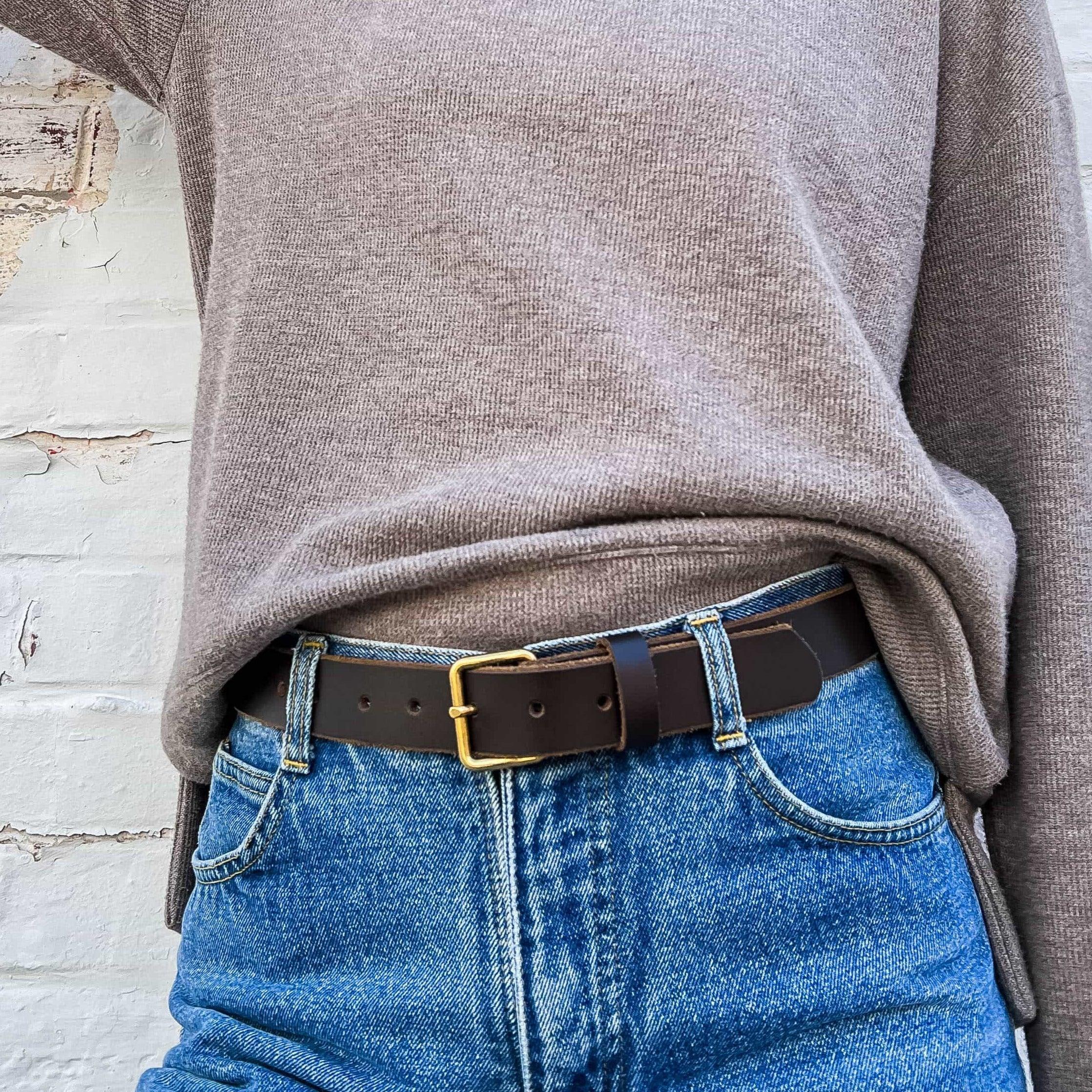 Brown leather belt with gold buckle on blue jeans and taupe sweater, white brick background