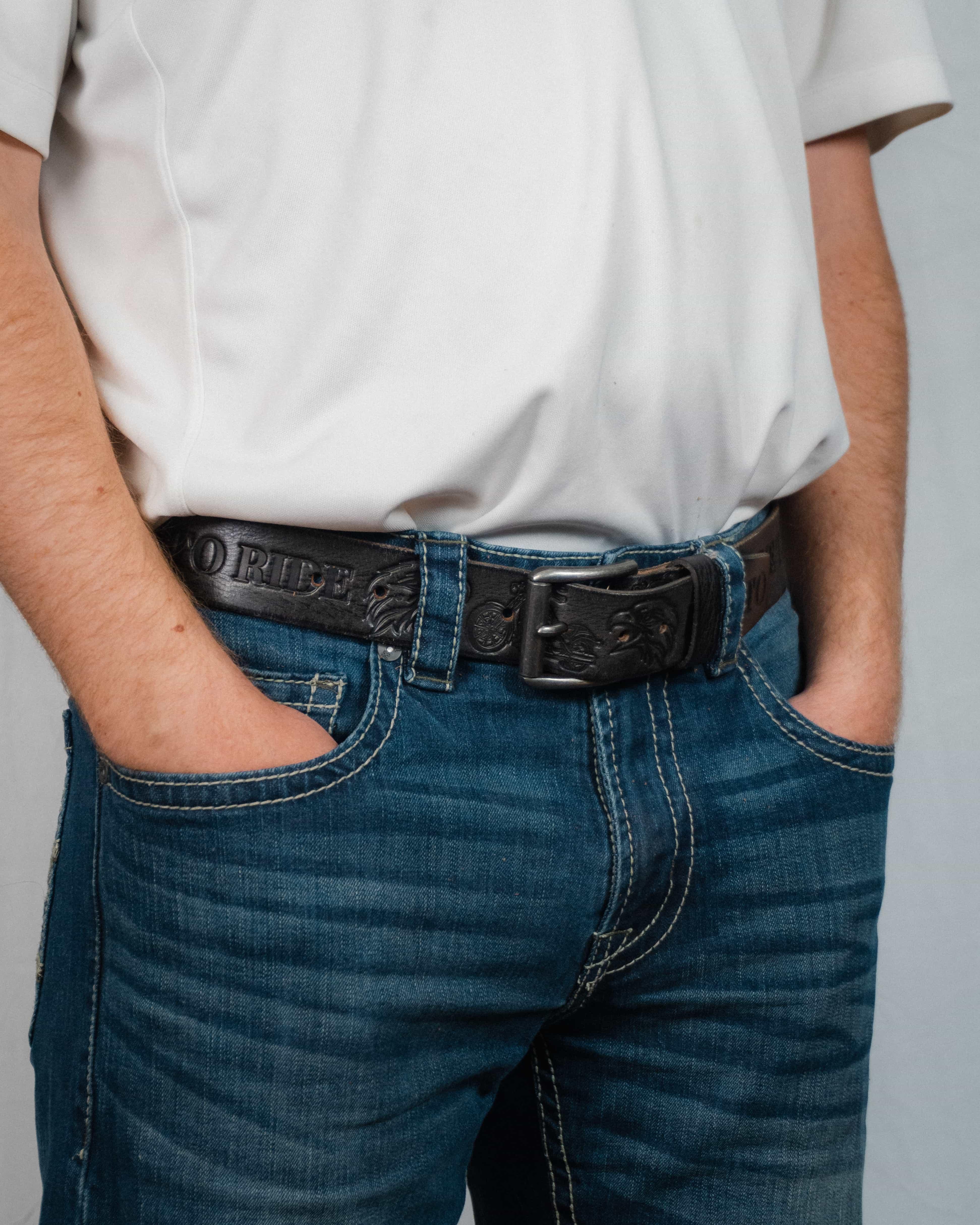 Man wearing blue jeans, white shirt, and black embossed leather belt with hands in pockets