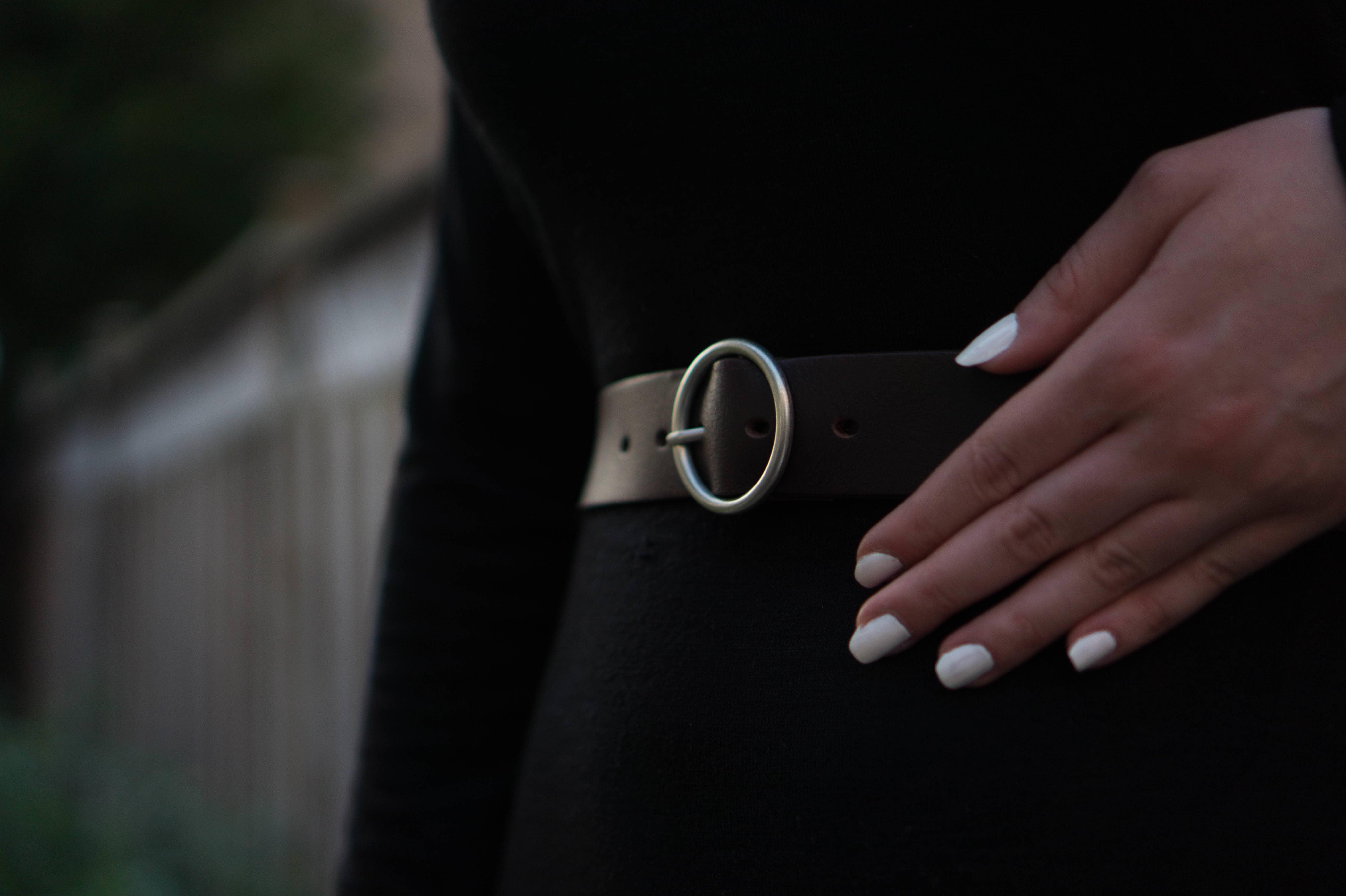 Brown leather belt with silver oval buckle on black dress, woman's hand with white nails