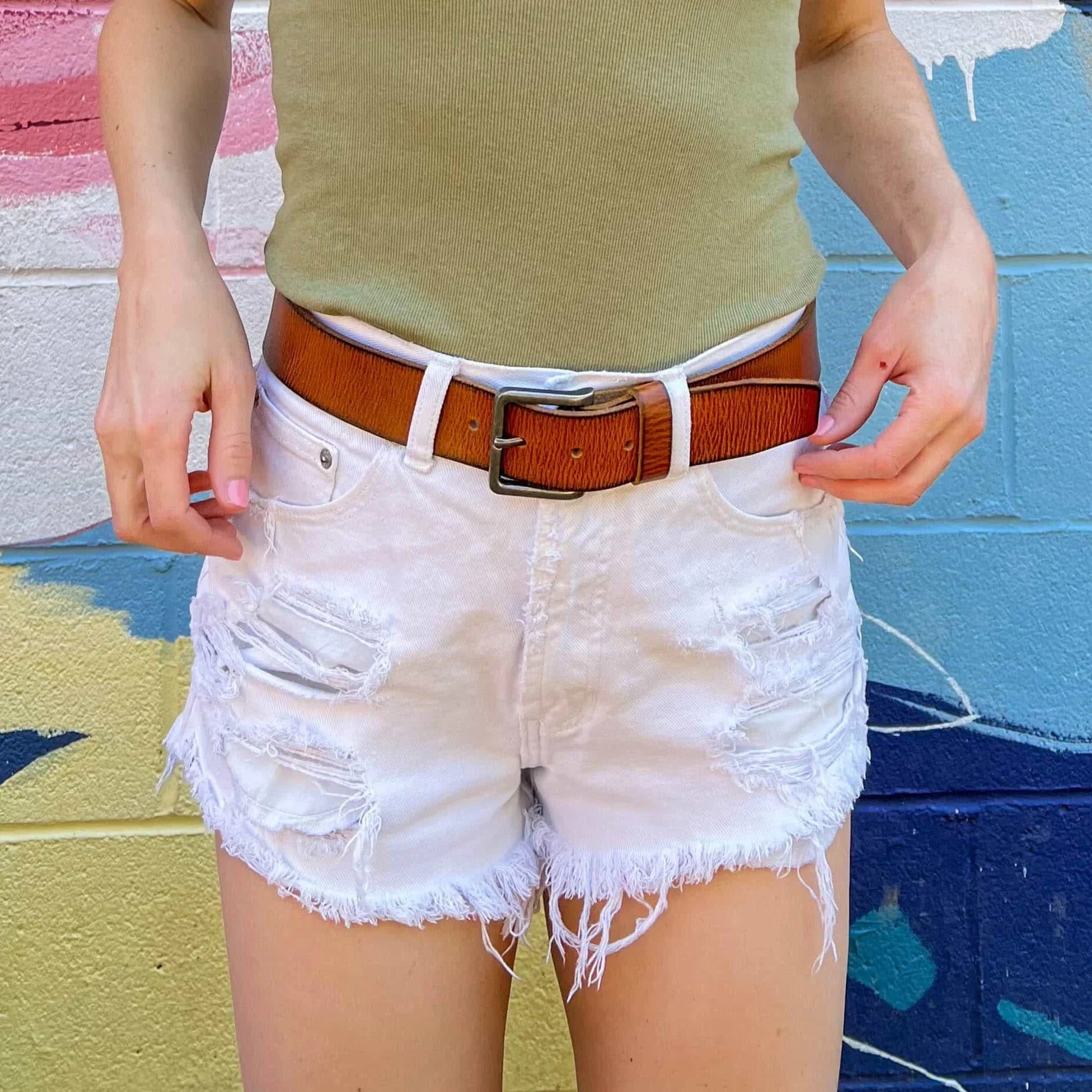 Woman wearing distressed white denim shorts and brown belt against colorful mural wall