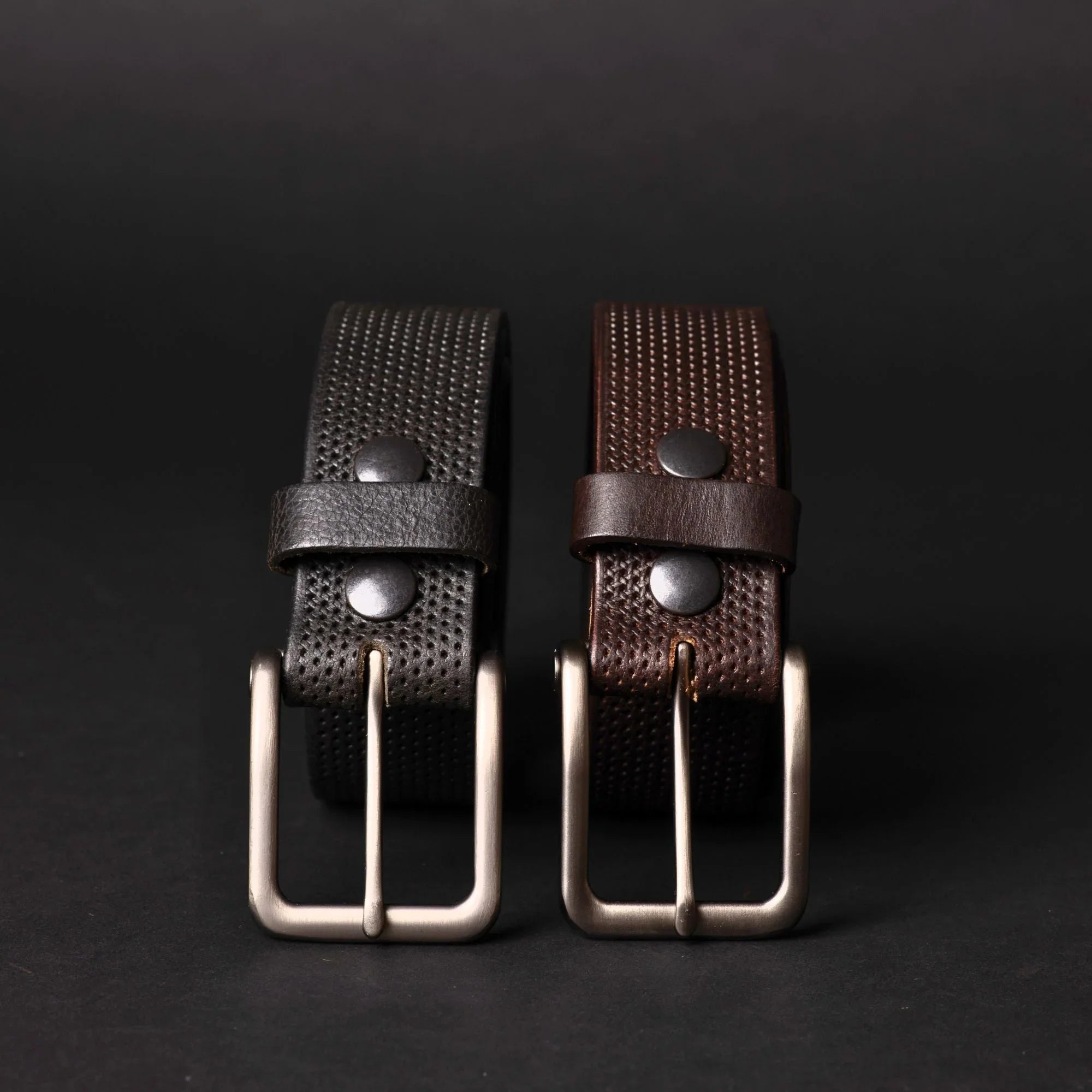 Two textured leather belts, black and brown, with metal buckles on a dark background.