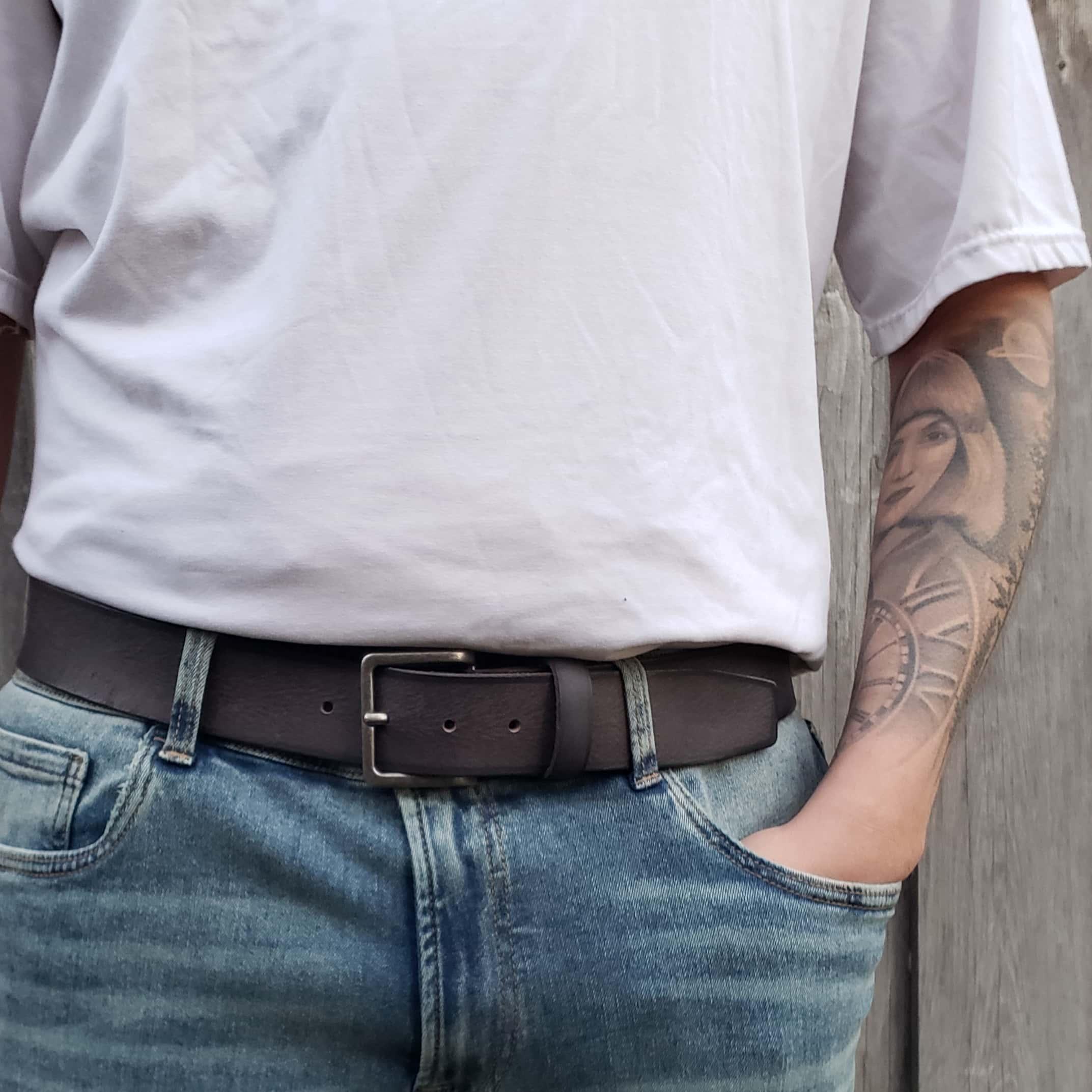 Man wearing white t-shirt, blue jeans, black leather belt, with tattooed arm outdoors