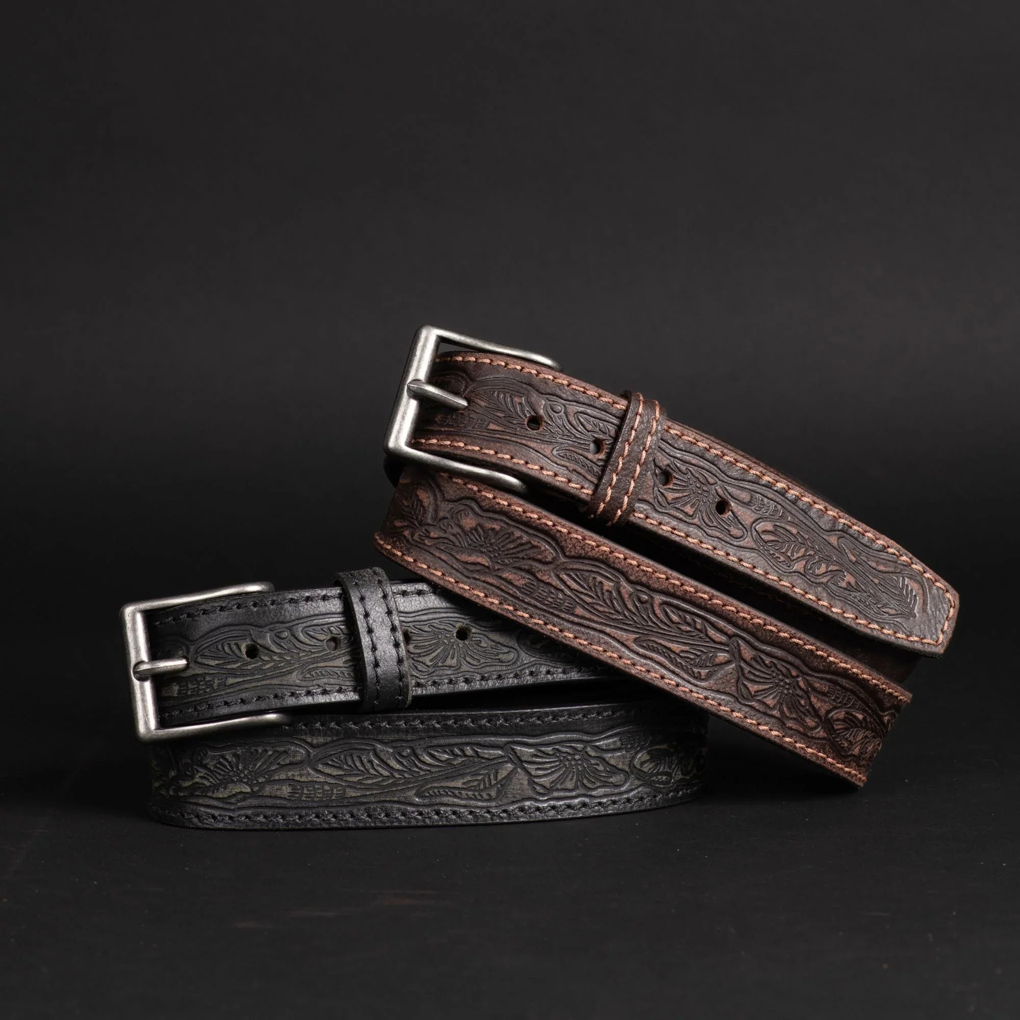 Black and brown embossed leather belts with floral patterns and metal buckles on black background.