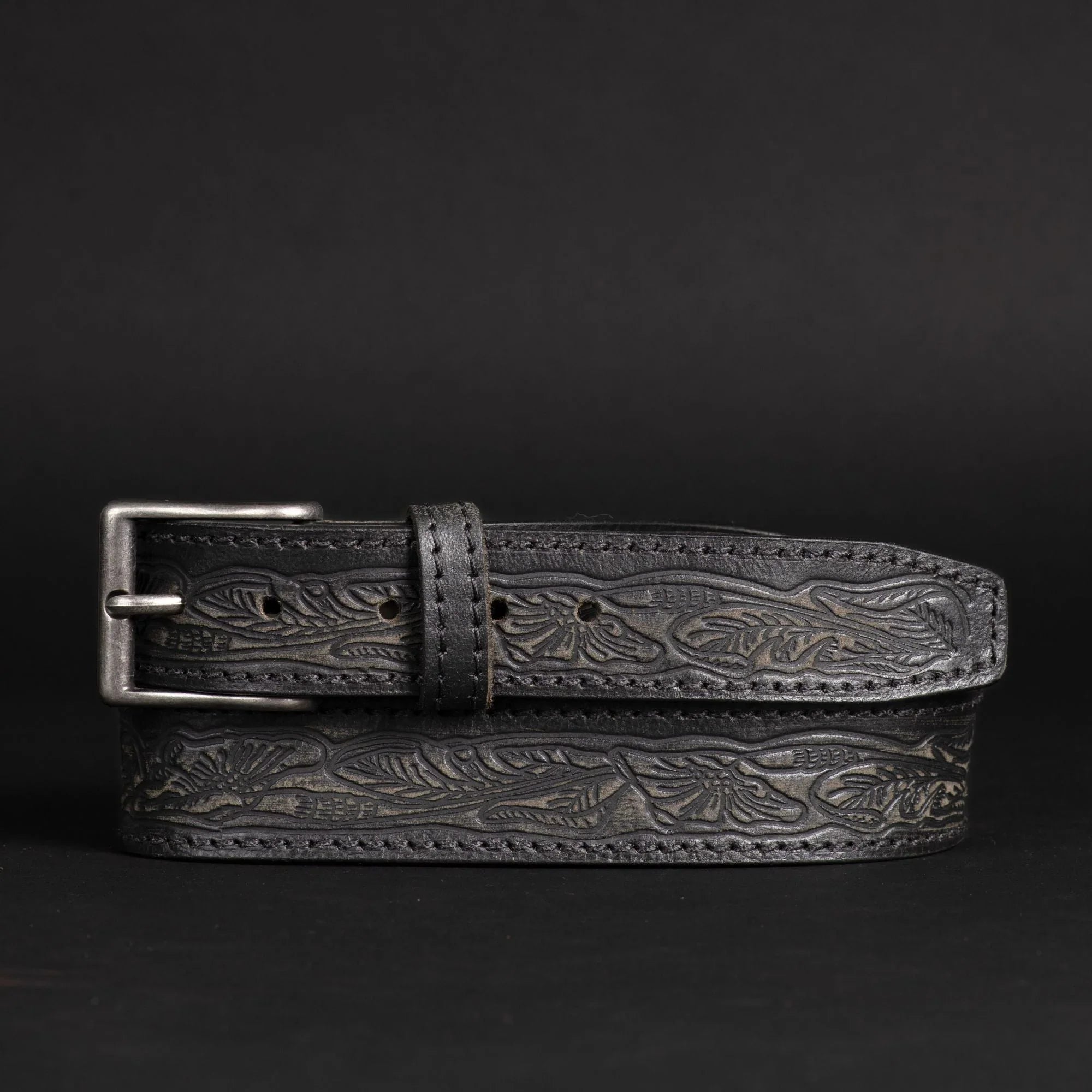 Black embossed leather belt with floral pattern and silver buckle on dark background