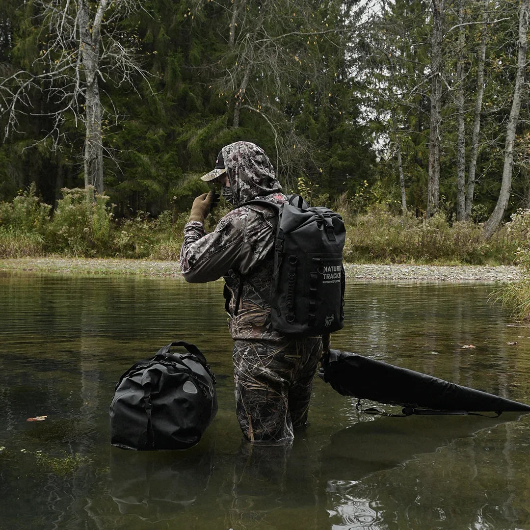 Hunter in camouflage gear with waterproof backpack and gear bag wading in forest stream