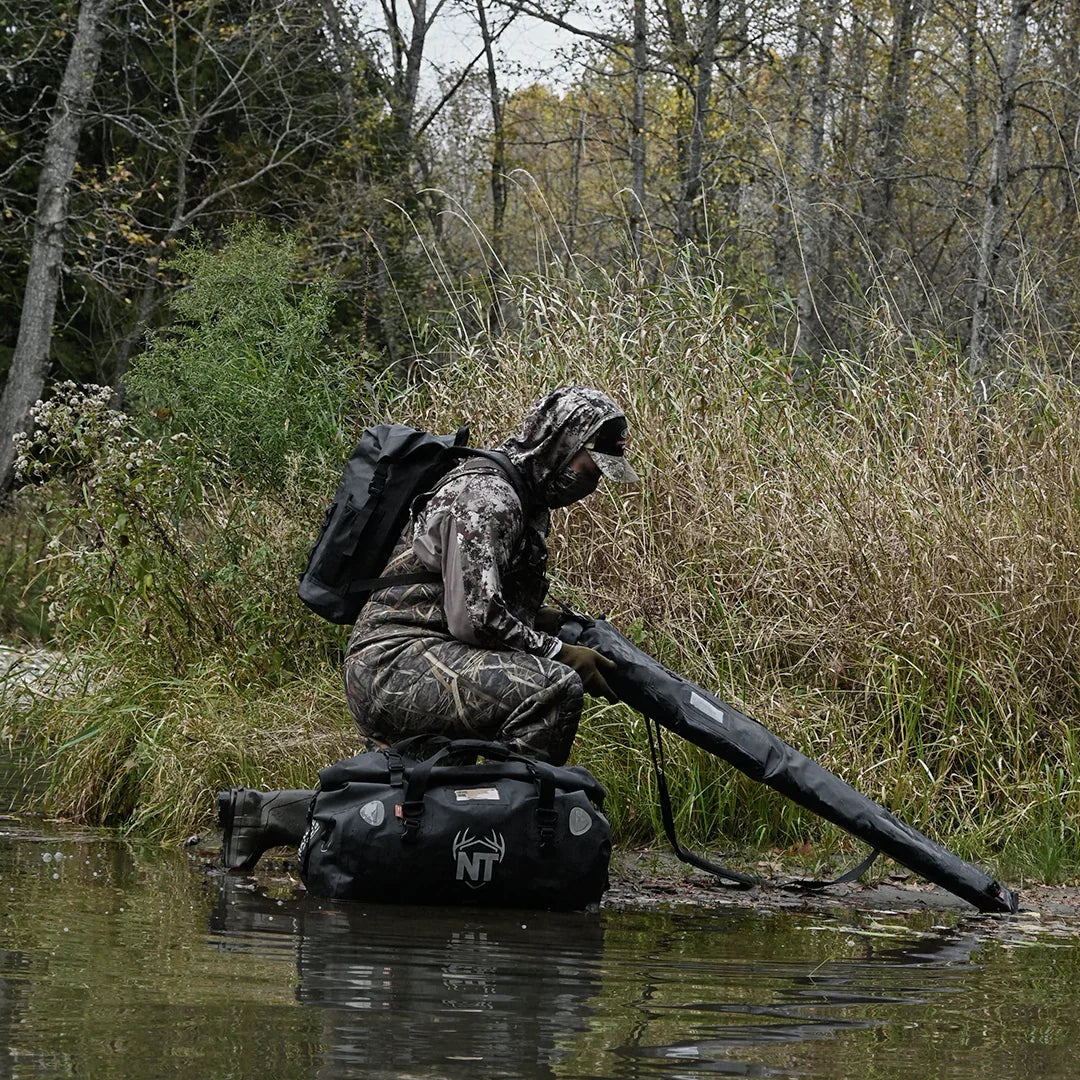 Hunter in camouflage gear with backpack and gun case by forest stream, hunting gear bag visible