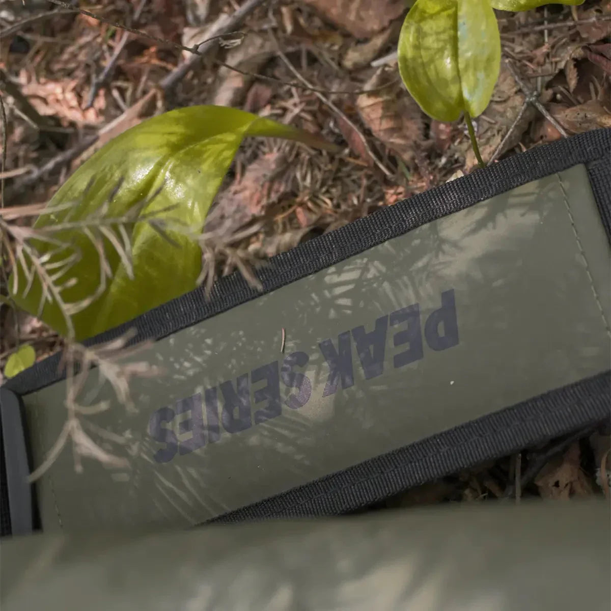 Olive green PEAK SERIES case on forest floor with green leaves and branches