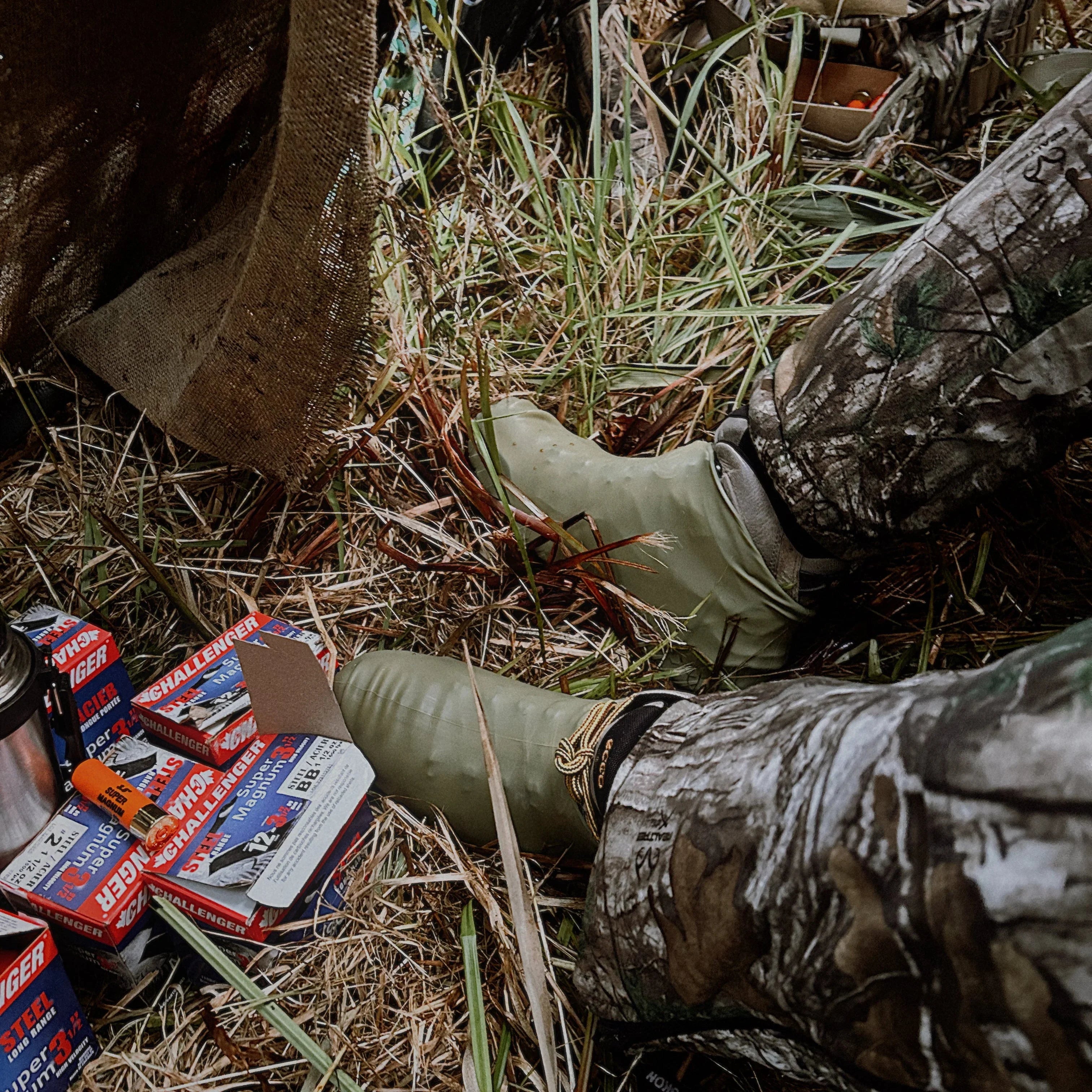 Green waterproof shoe covers on camo pants, shotgun shell boxes on grass outdoors