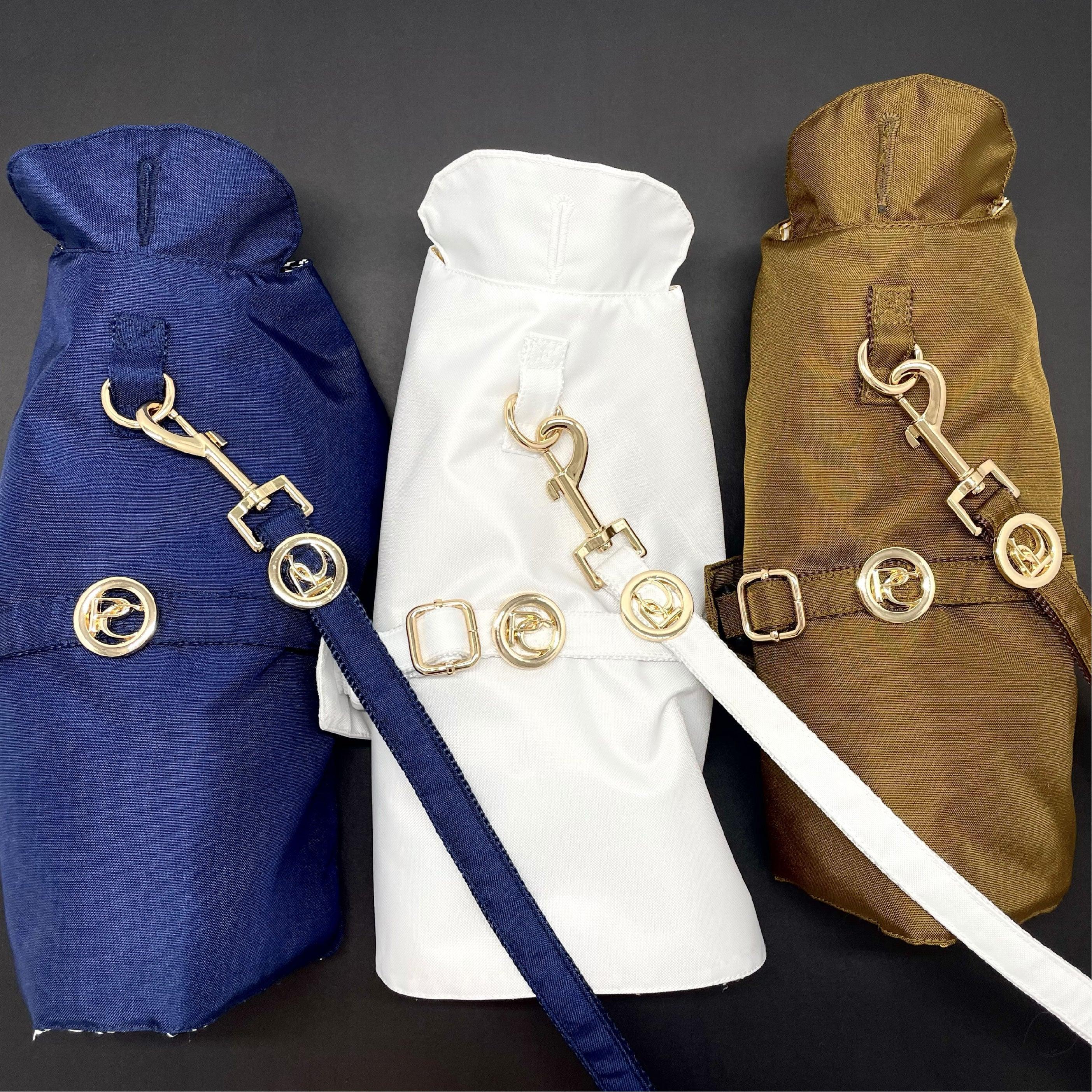 Three stylish dog raincoats in navy, white, and brown with gold hardware on black background