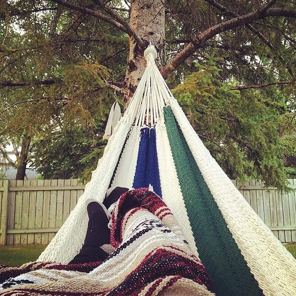 Blue, green, and white Nicaraguan hammock hanging in a backyard with trees and fence.