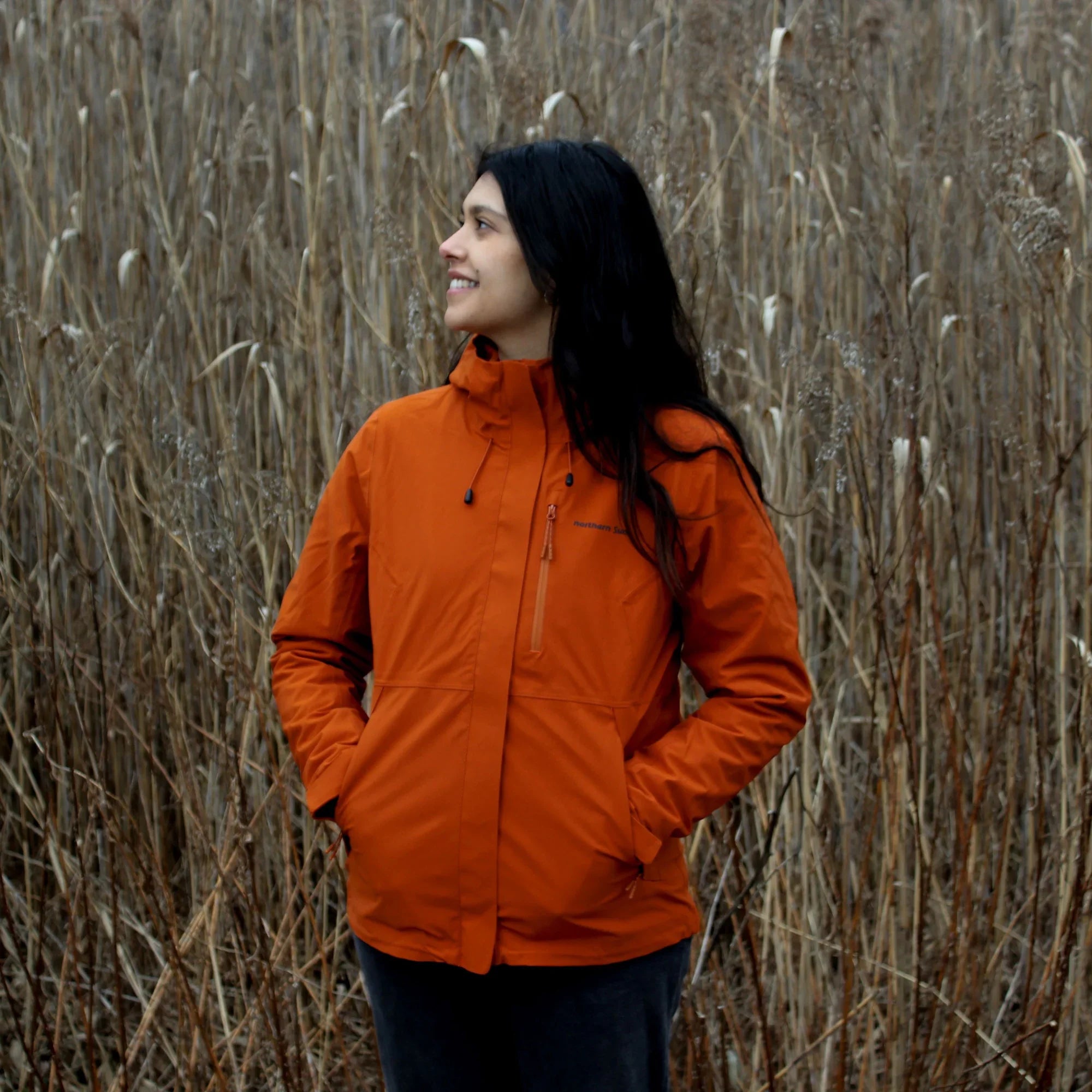 Woman wearing orange waterproof fall jacket standing in tall dry grass outdoors