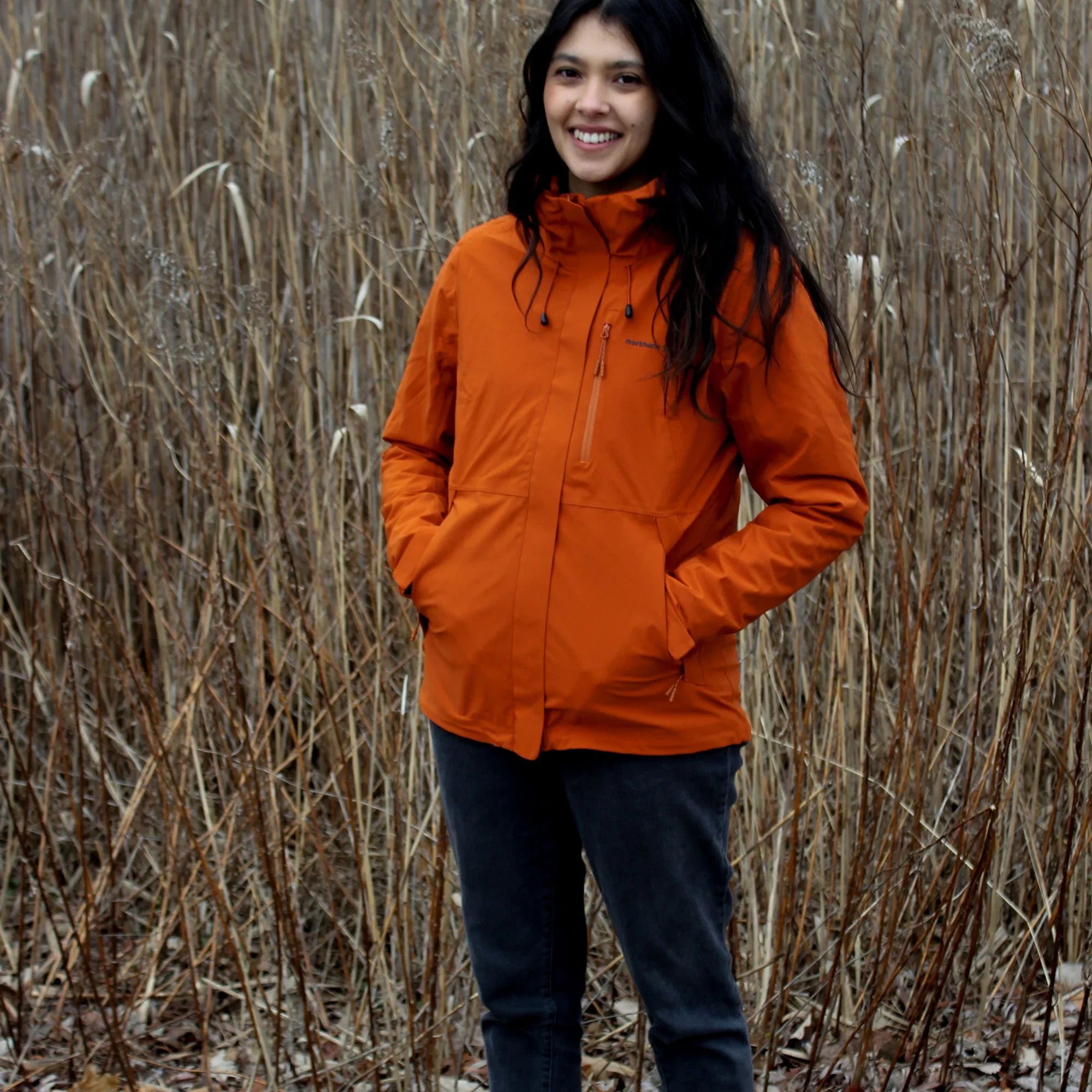 Woman in orange 3-in-1 waterproof winter jacket standing outdoors in tall dry grass