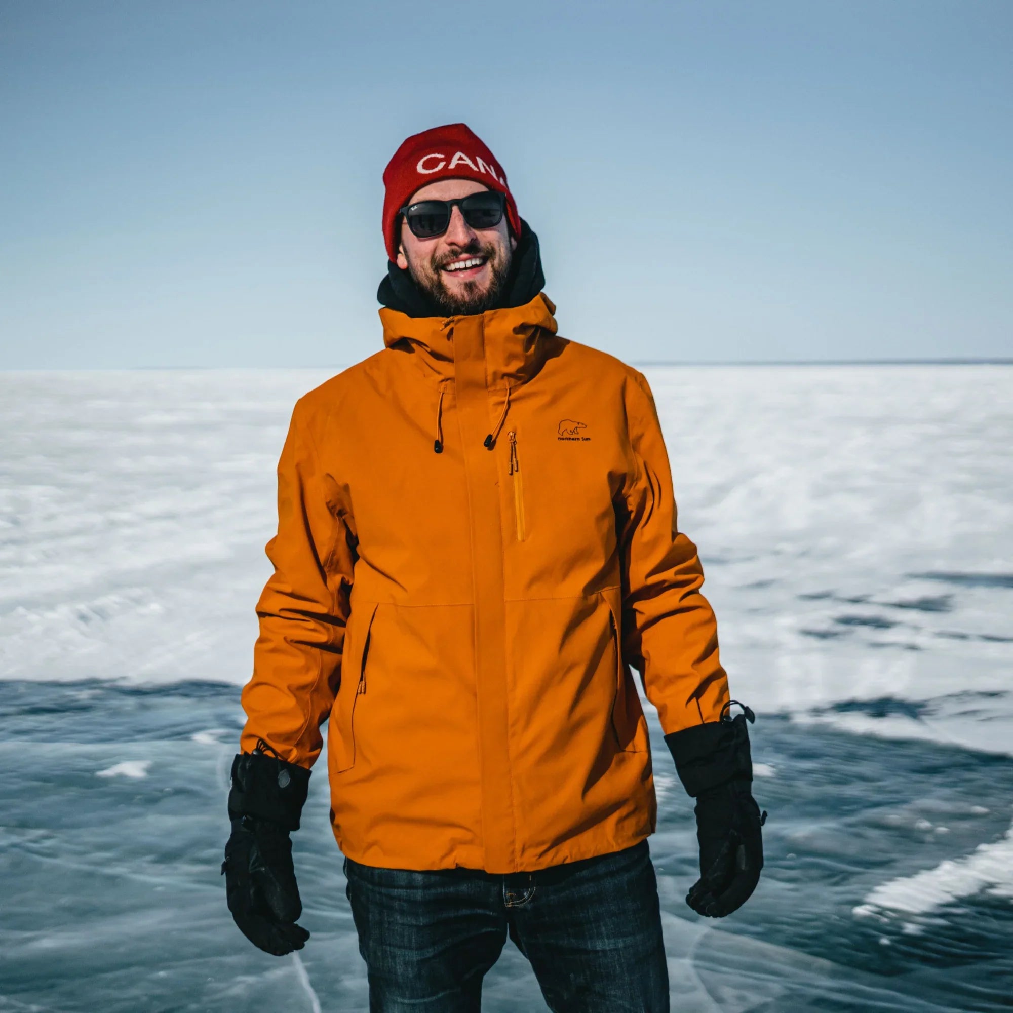 Man wearing orange 3-in-1 winter jacket on snow-covered landscape, sunglasses, red beanie