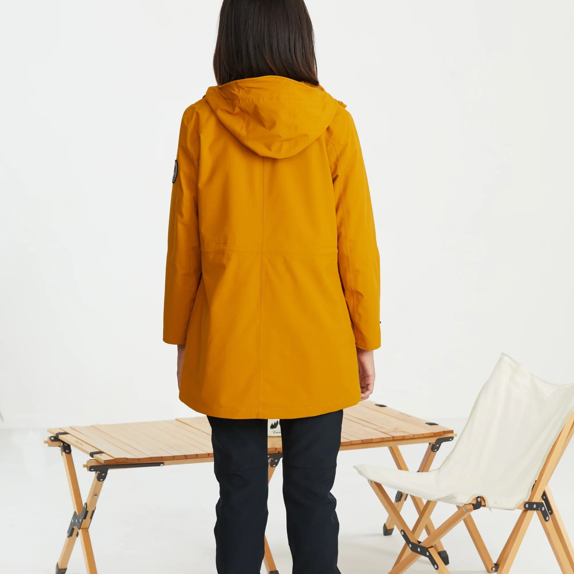 Woman wearing yellow mid-length 3-in-1 jacket, standing by camping table and chair indoors