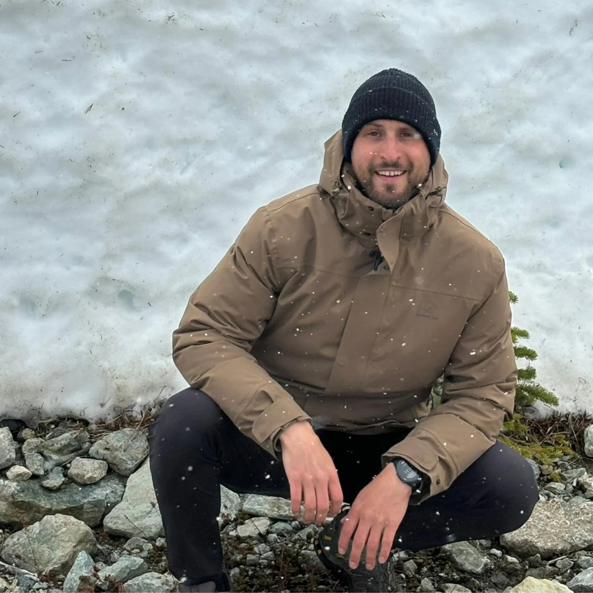 Man in brown winter jacket and black beanie posing outdoors on snowy mountain terrain
