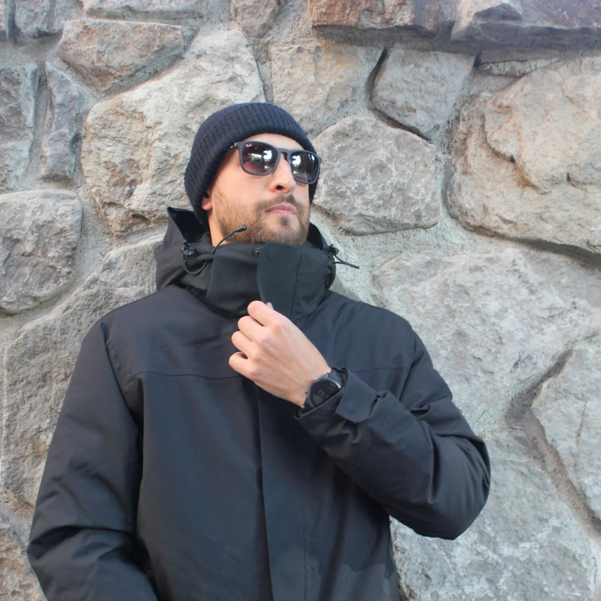 Man in black winter jacket, beanie, and sunglasses standing by stone wall