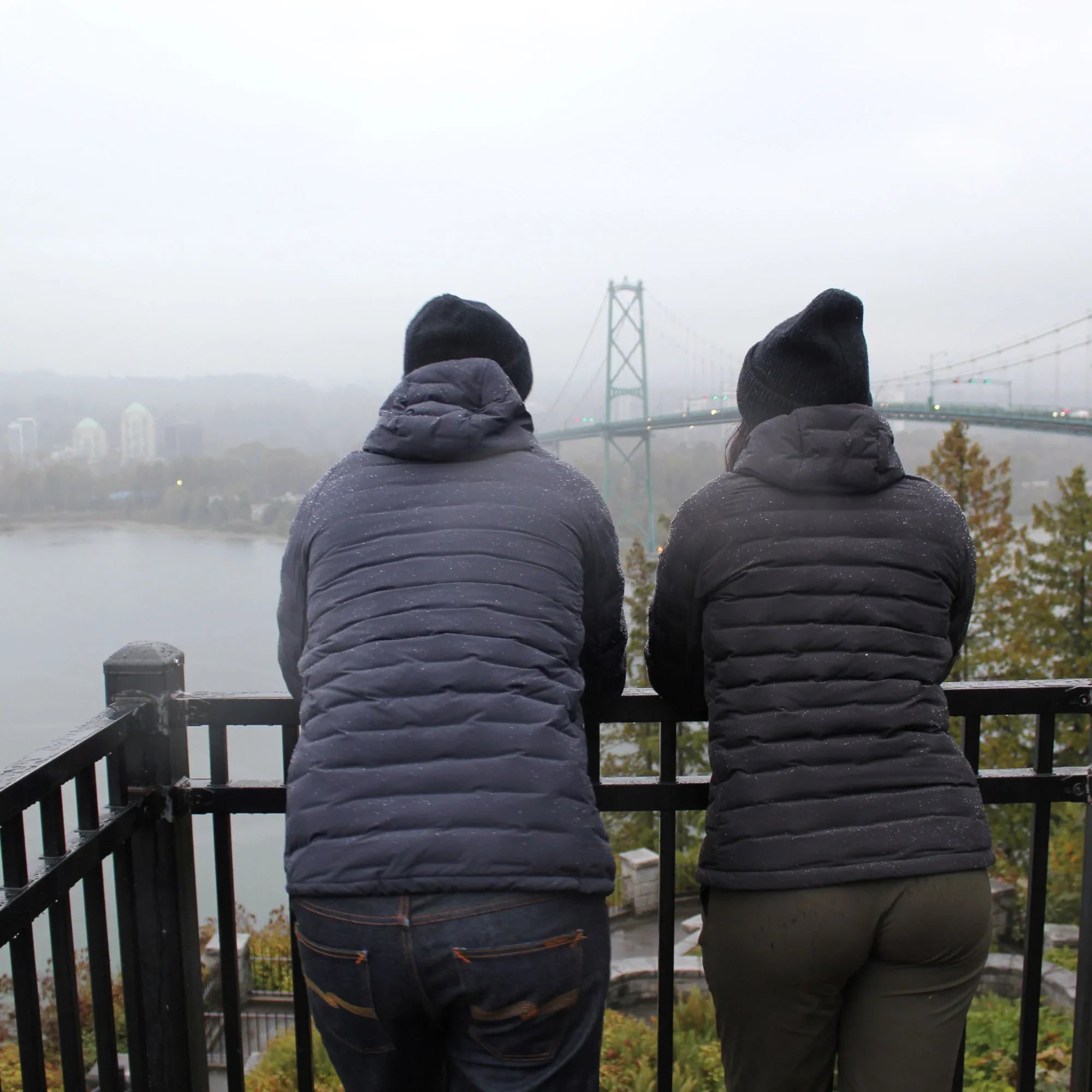 Two people in hooded down jackets and beanies facing a foggy bridge view outdoors