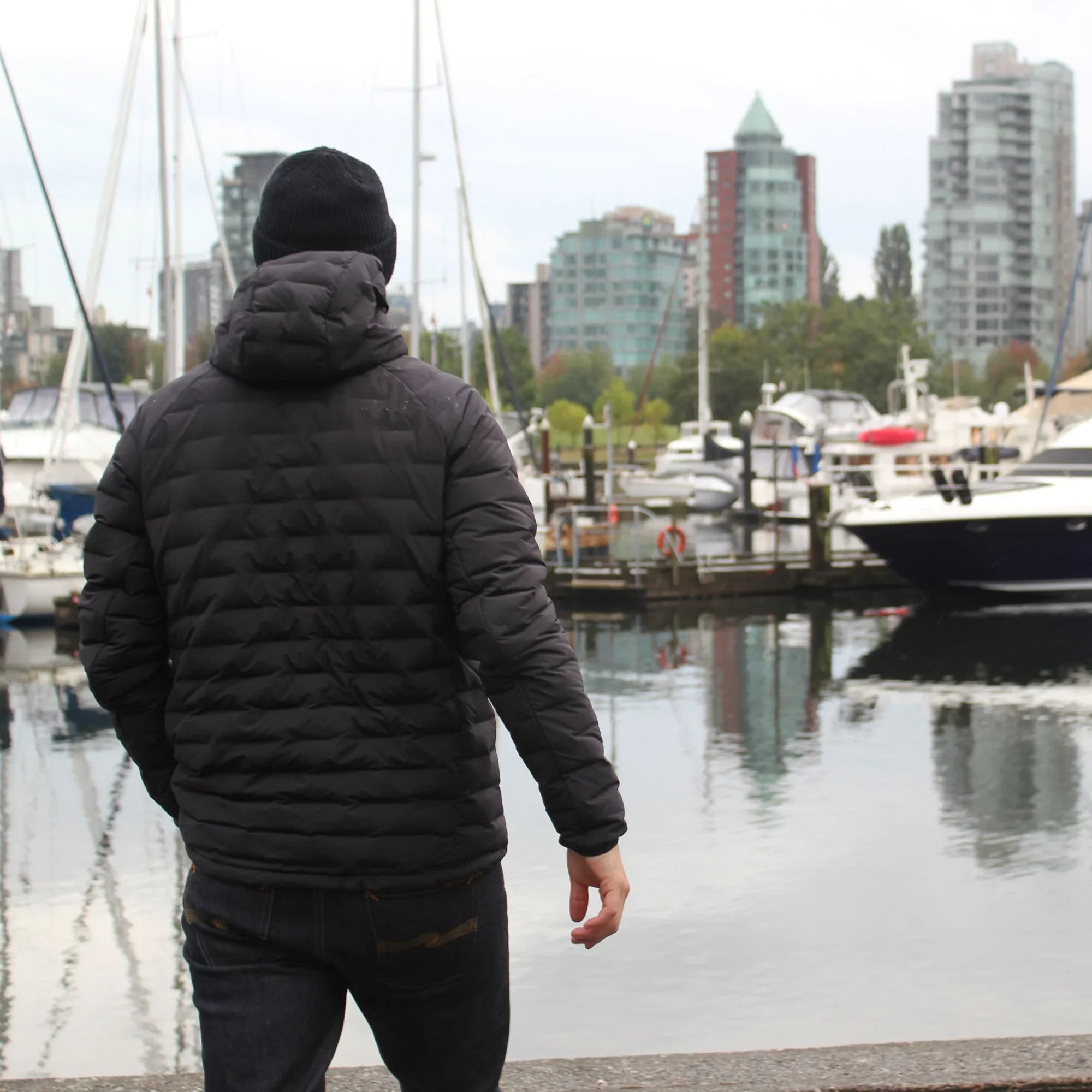 Man wearing black lightweight down jacket by Vancouver marina with boats and cityscape
