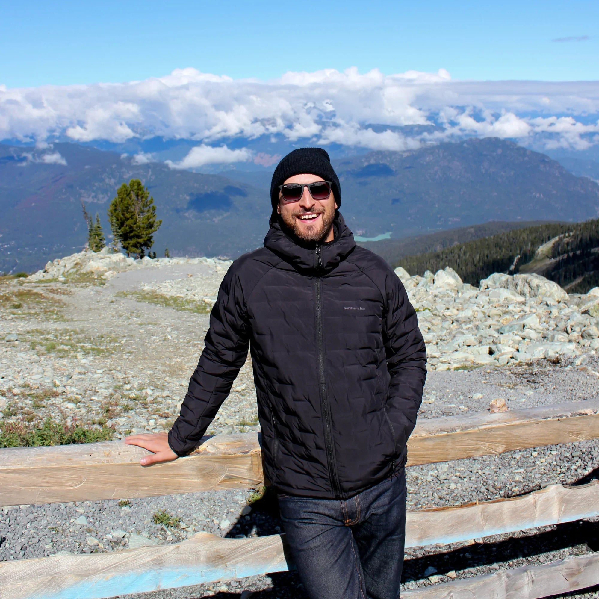 Man wearing black down puffer jacket and beanie with mountains and blue sky in background