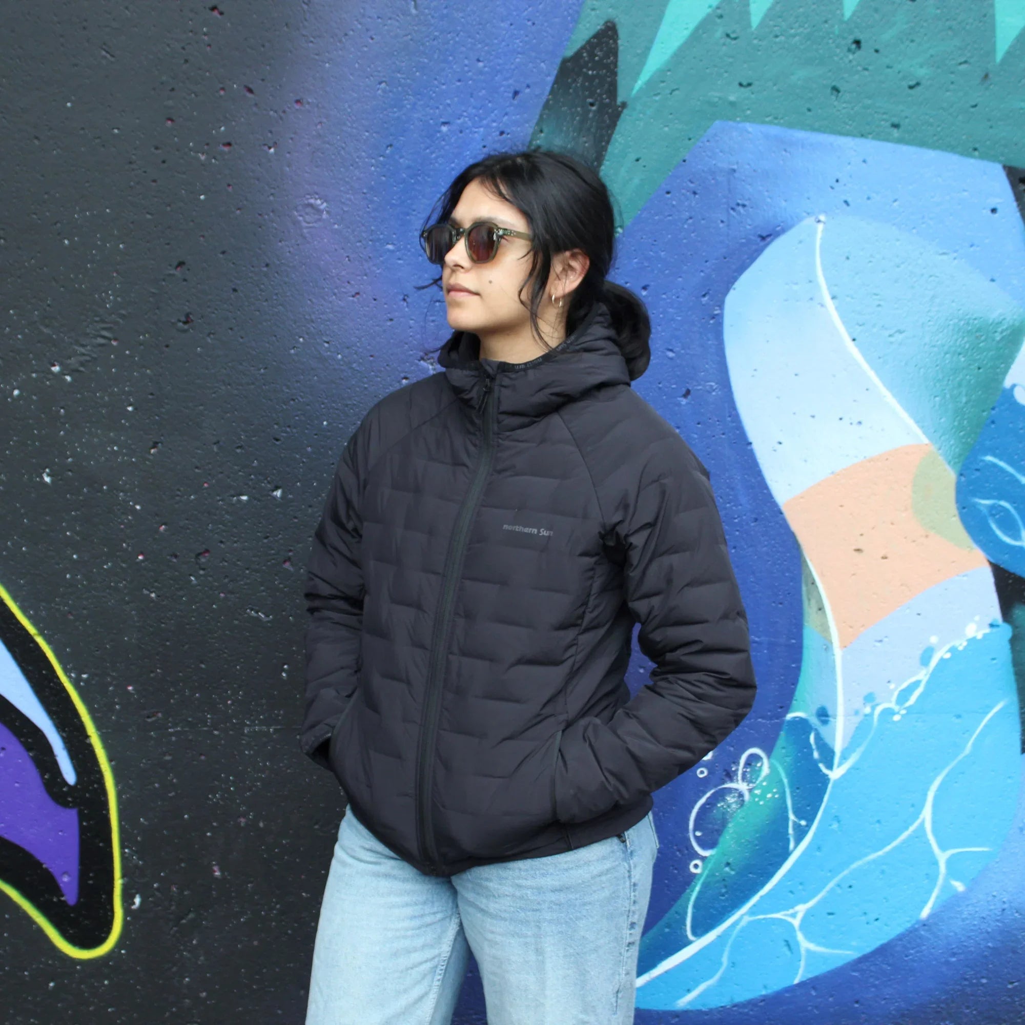 Woman in black lightweight down jacket and jeans standing by colorful urban mural