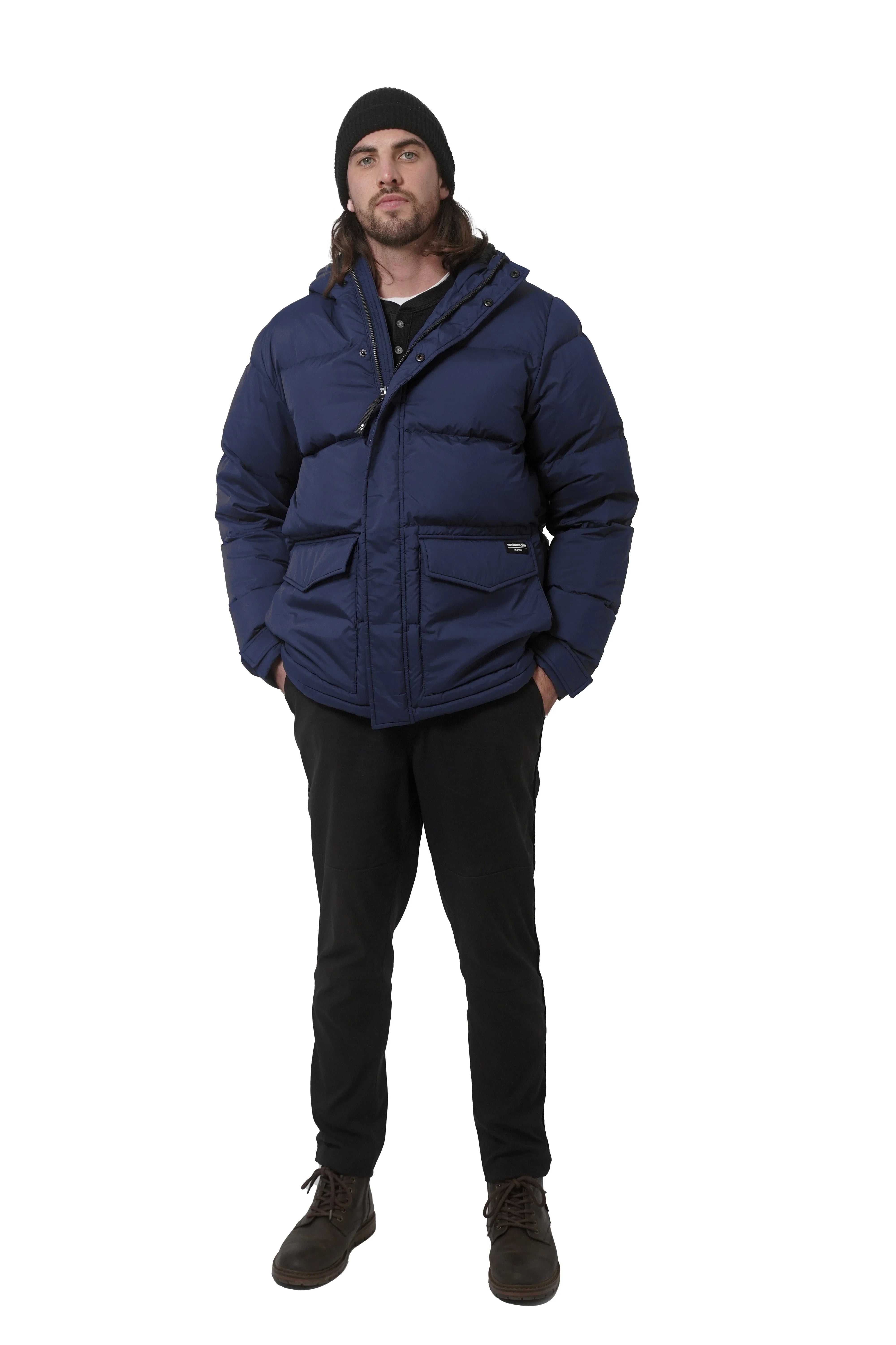Man wearing navy blue winter down parka, black beanie, black pants, brown boots, studio background