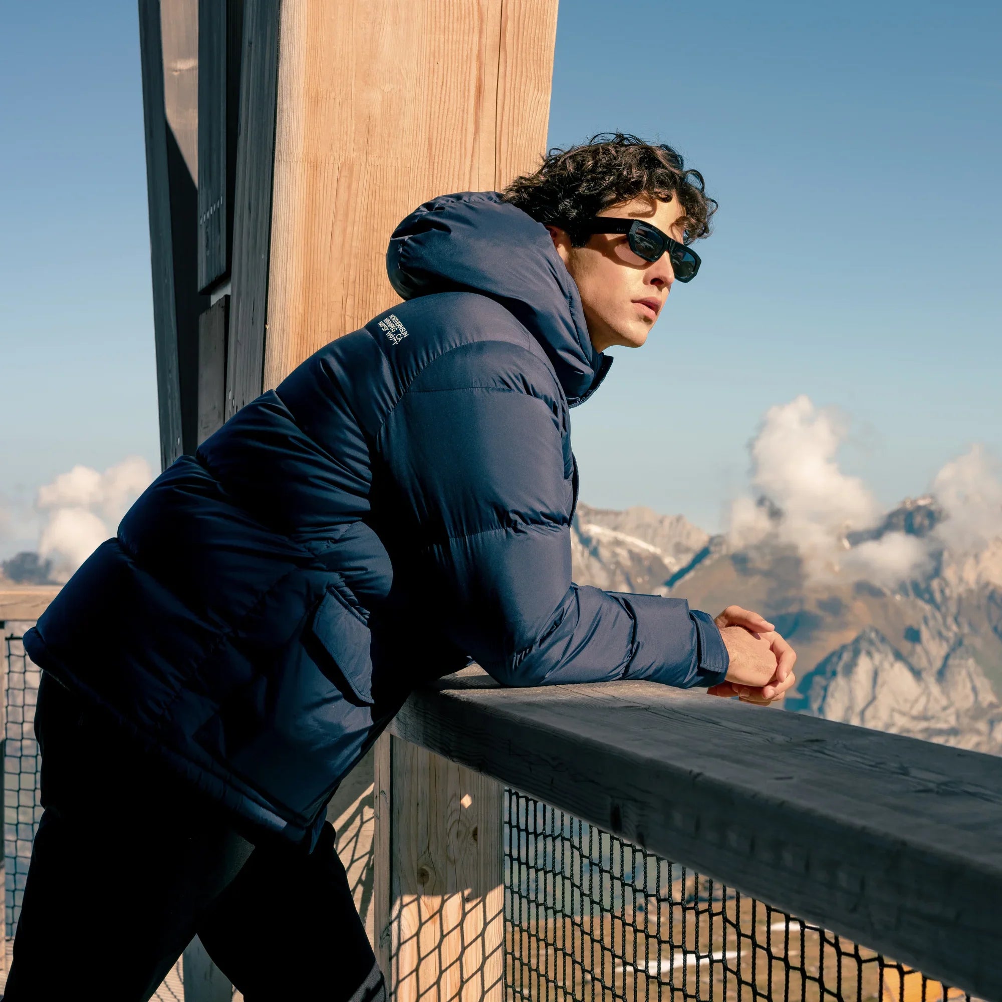 Man in navy winter down parka and sunglasses overlooking mountains from wooden balcony