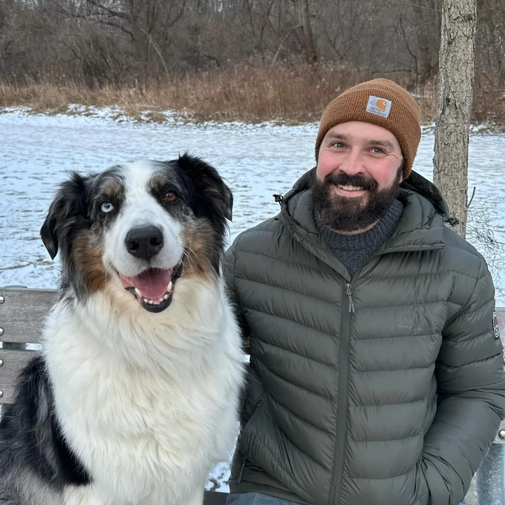 Man in olive lightweight down jacket and brown beanie with dog outdoors in snowy winter park