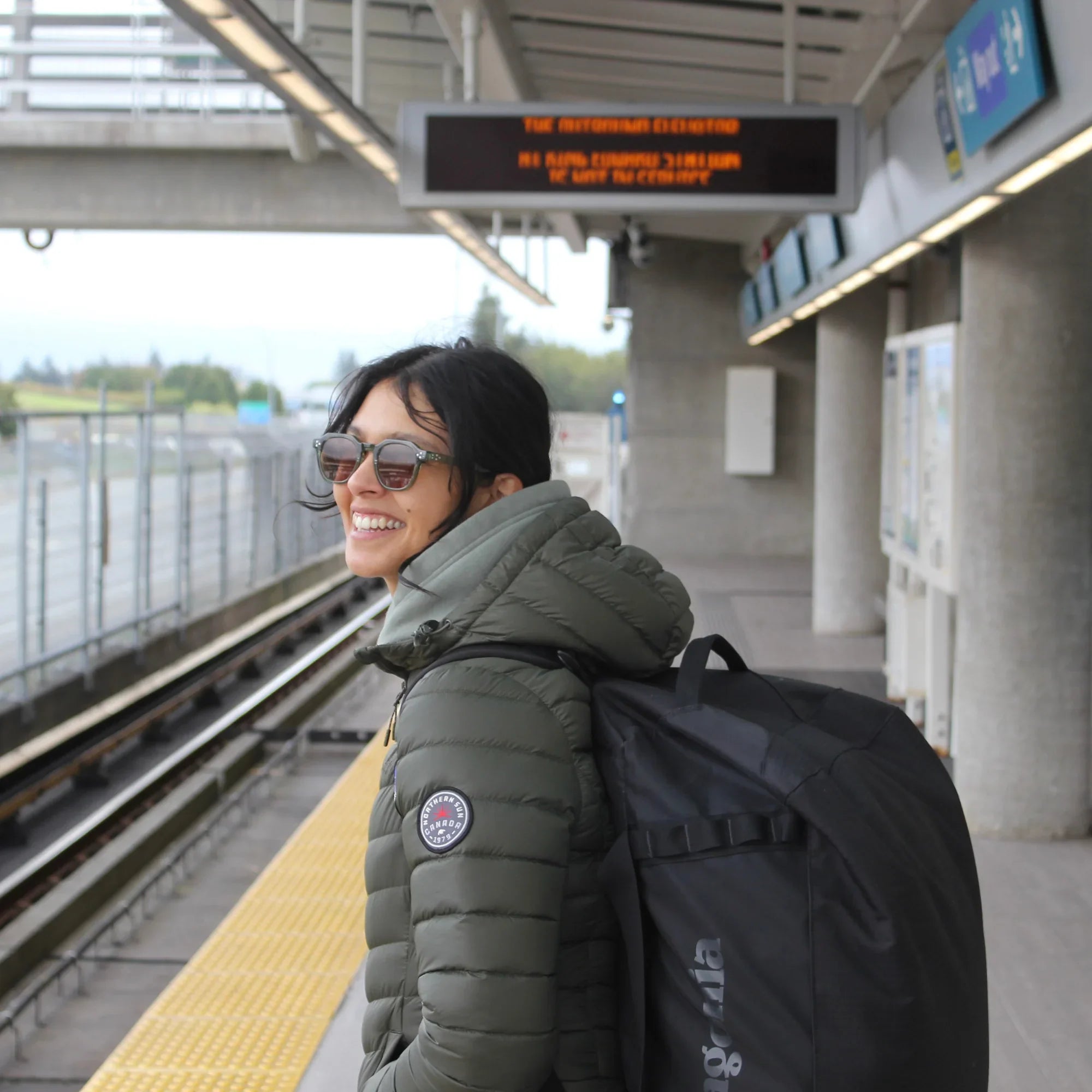 Woman in olive green packable lightweight down jacket waiting at sky train station with backpack