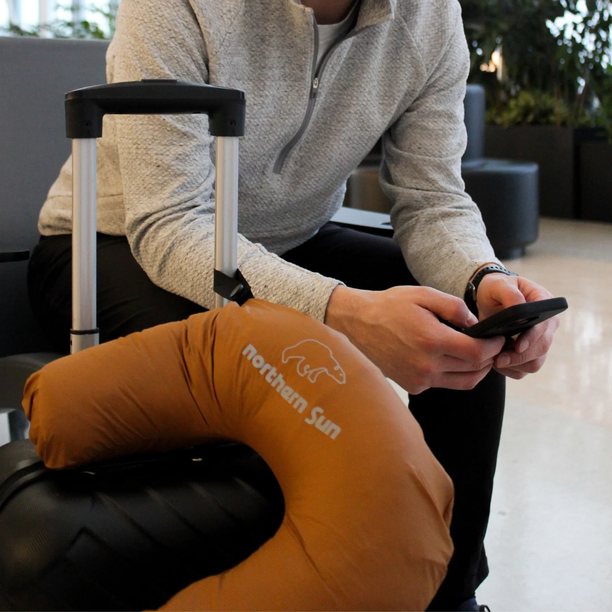 Man at airport with suitcase, using phone, Northern Sun neck pillow visible