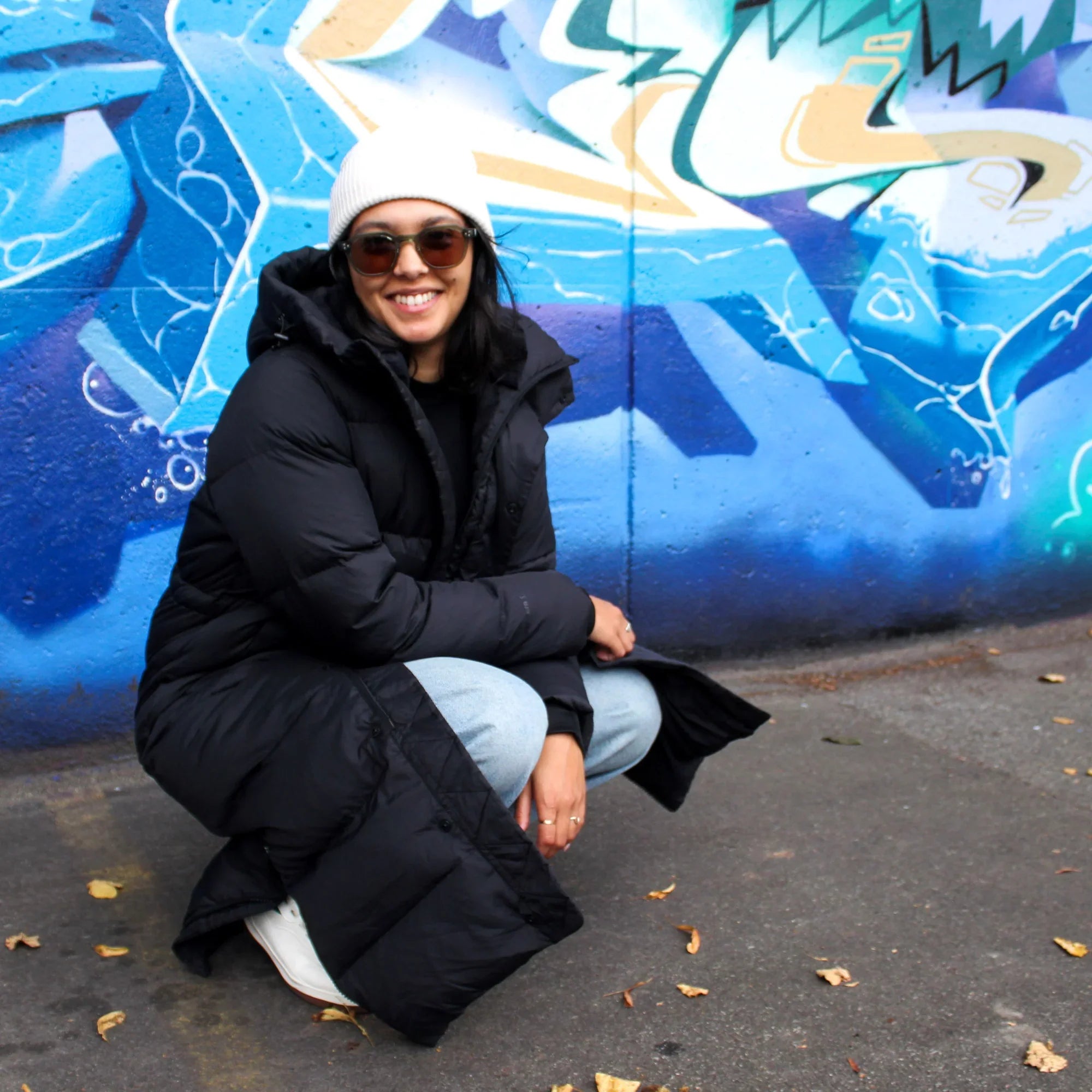 Woman in black puffer coat and white beanie smiling by blue graffiti wall
