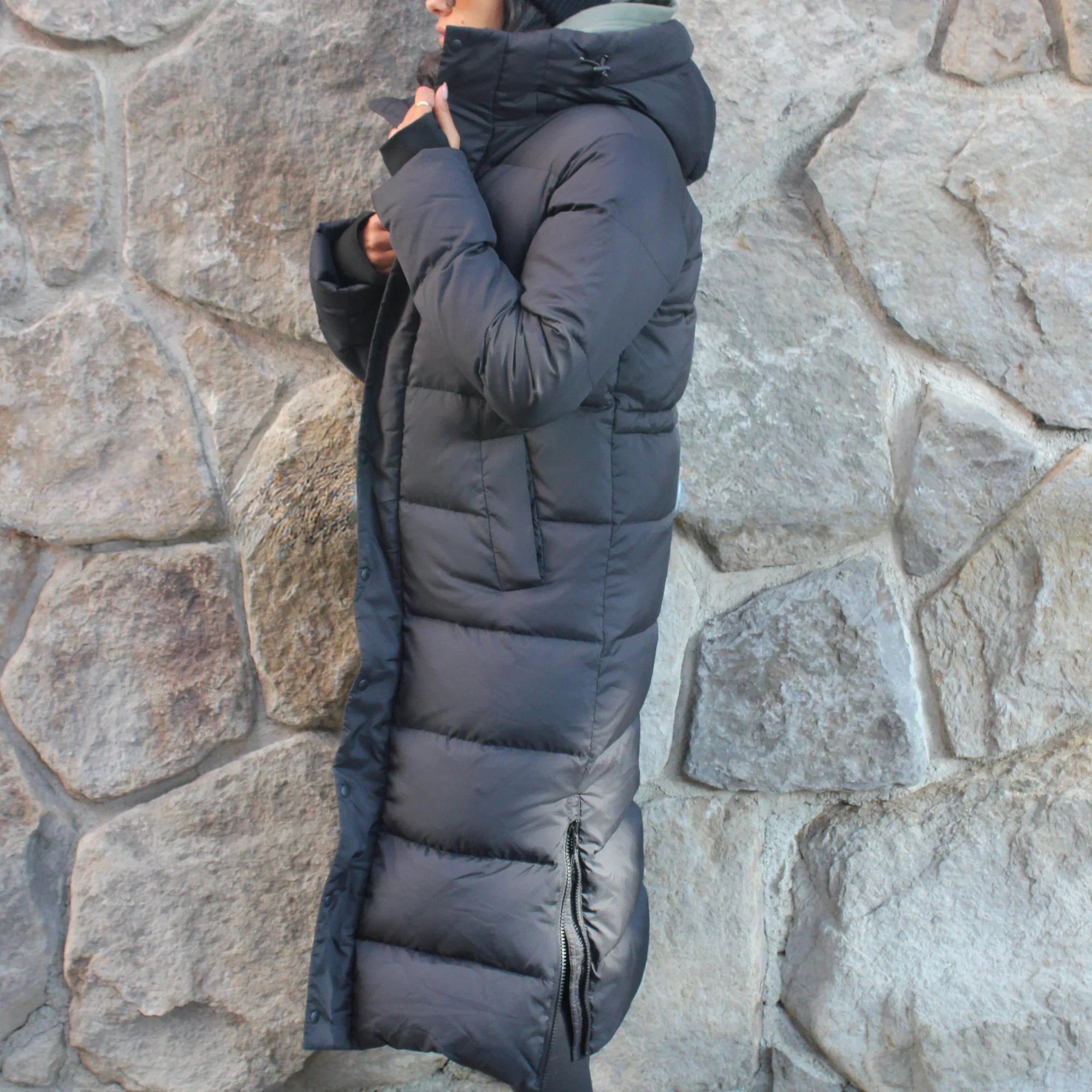 Woman in long black puffer winter coat with hood, standing by a stone wall