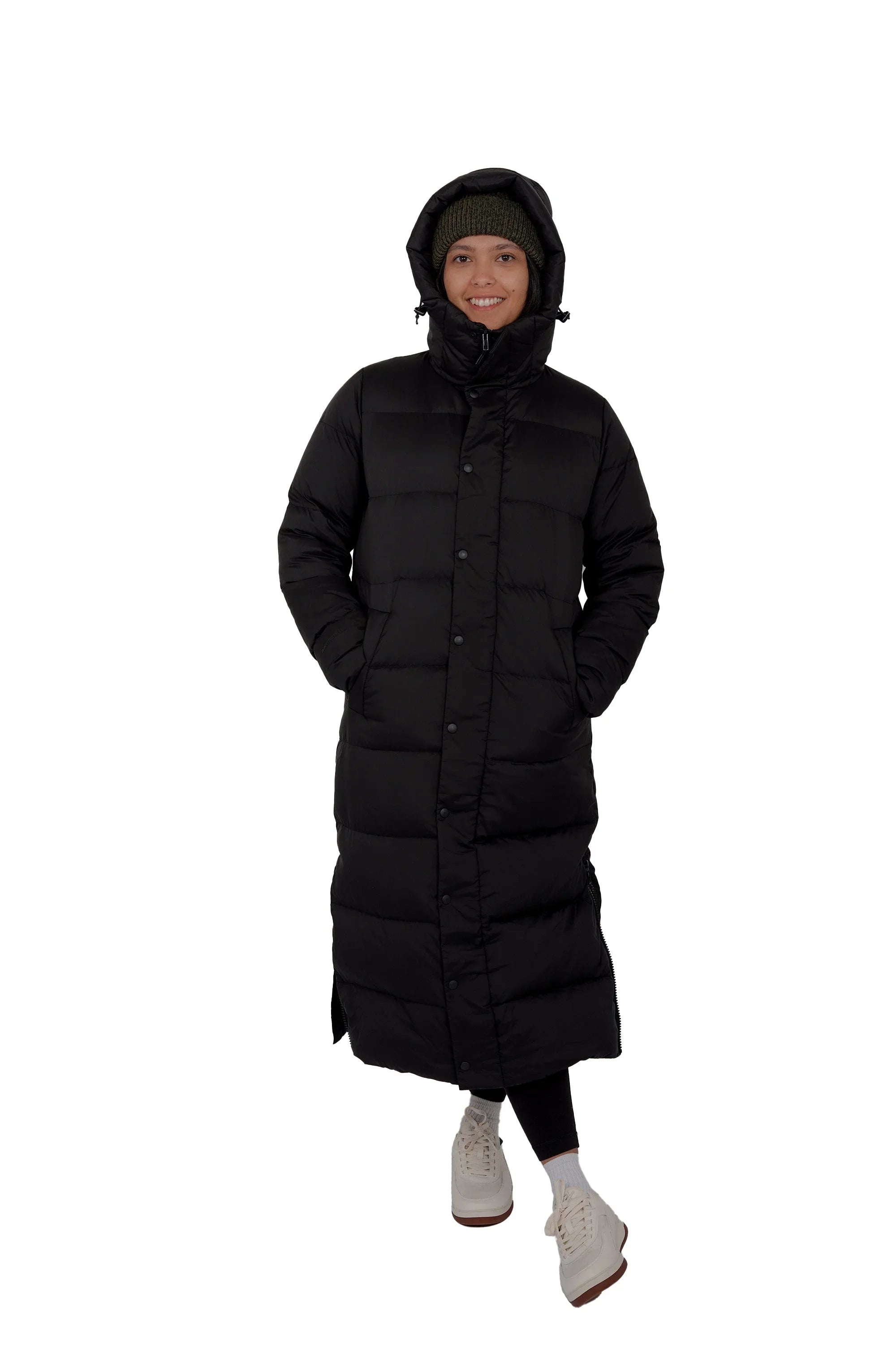 Woman in long black puffer coat with hood, green knit hat, and white sneakers on white background