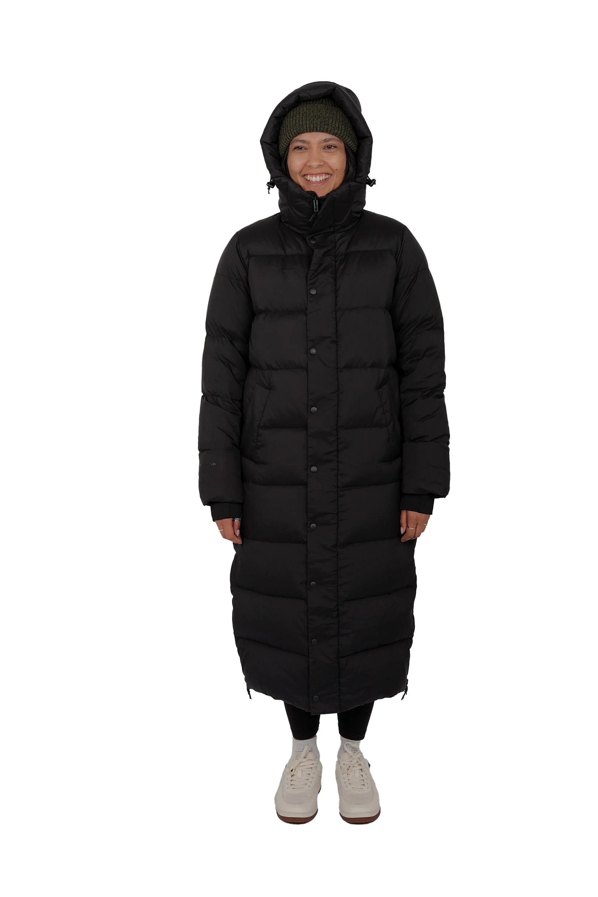 Woman wearing long black puffer coat with hood, green beanie, white sneakers