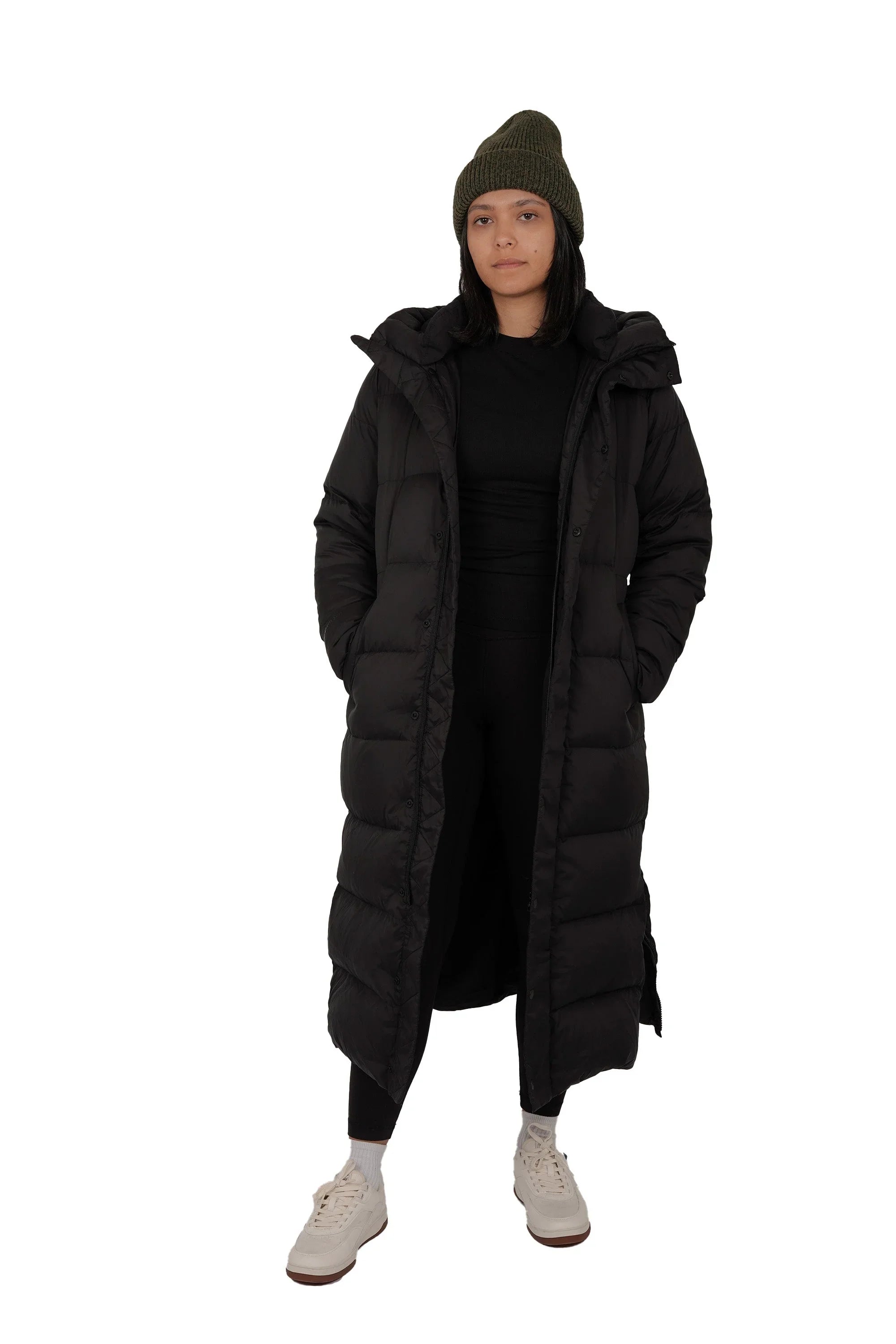 Woman in long black puffer coat, green knit beanie, white sneakers, on white background