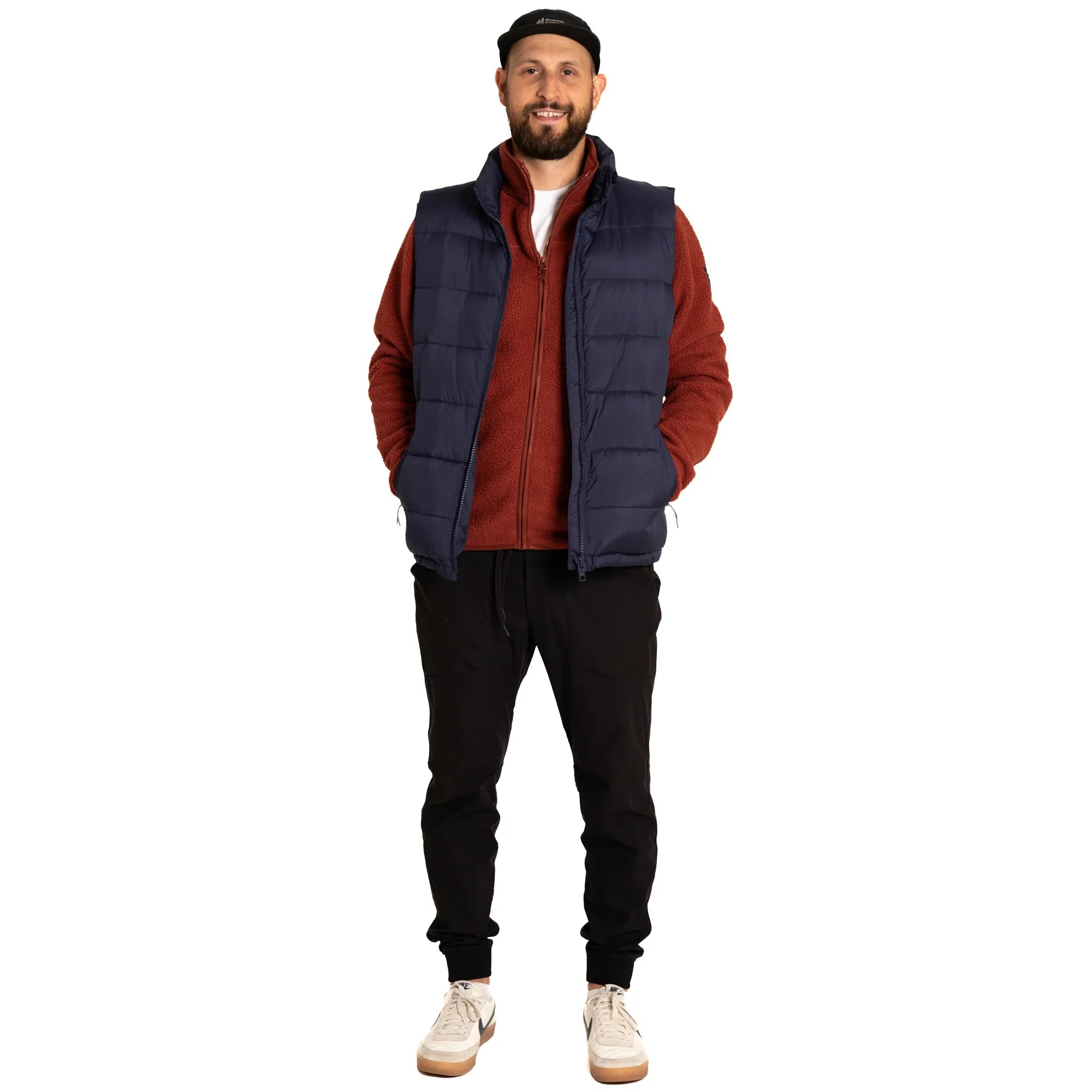 Man in navy puffer vest, red zip-up jacket, black joggers, white sneakers, and black cap