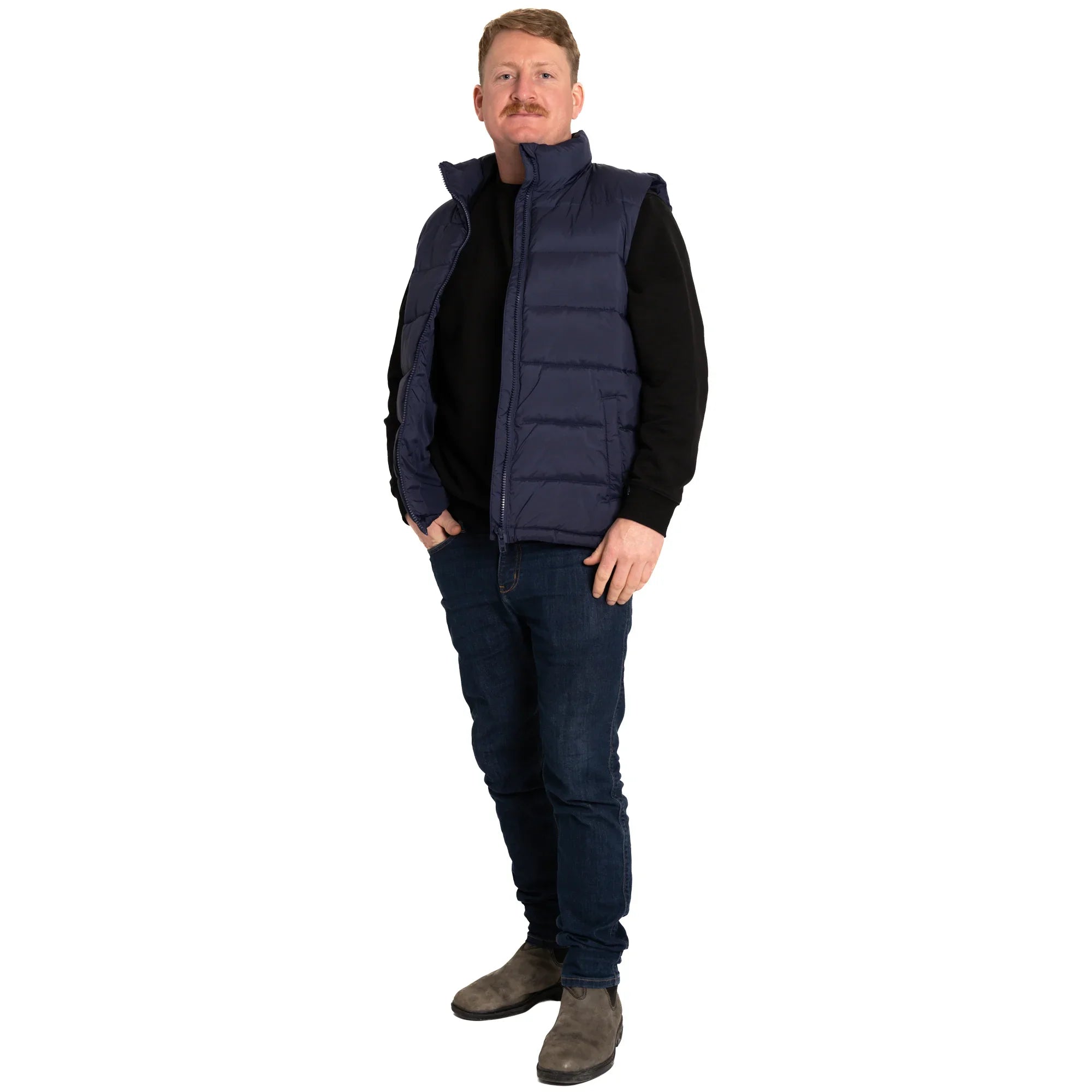 Man wearing navy puffer vest, black sweater, dark jeans, and boots on white background