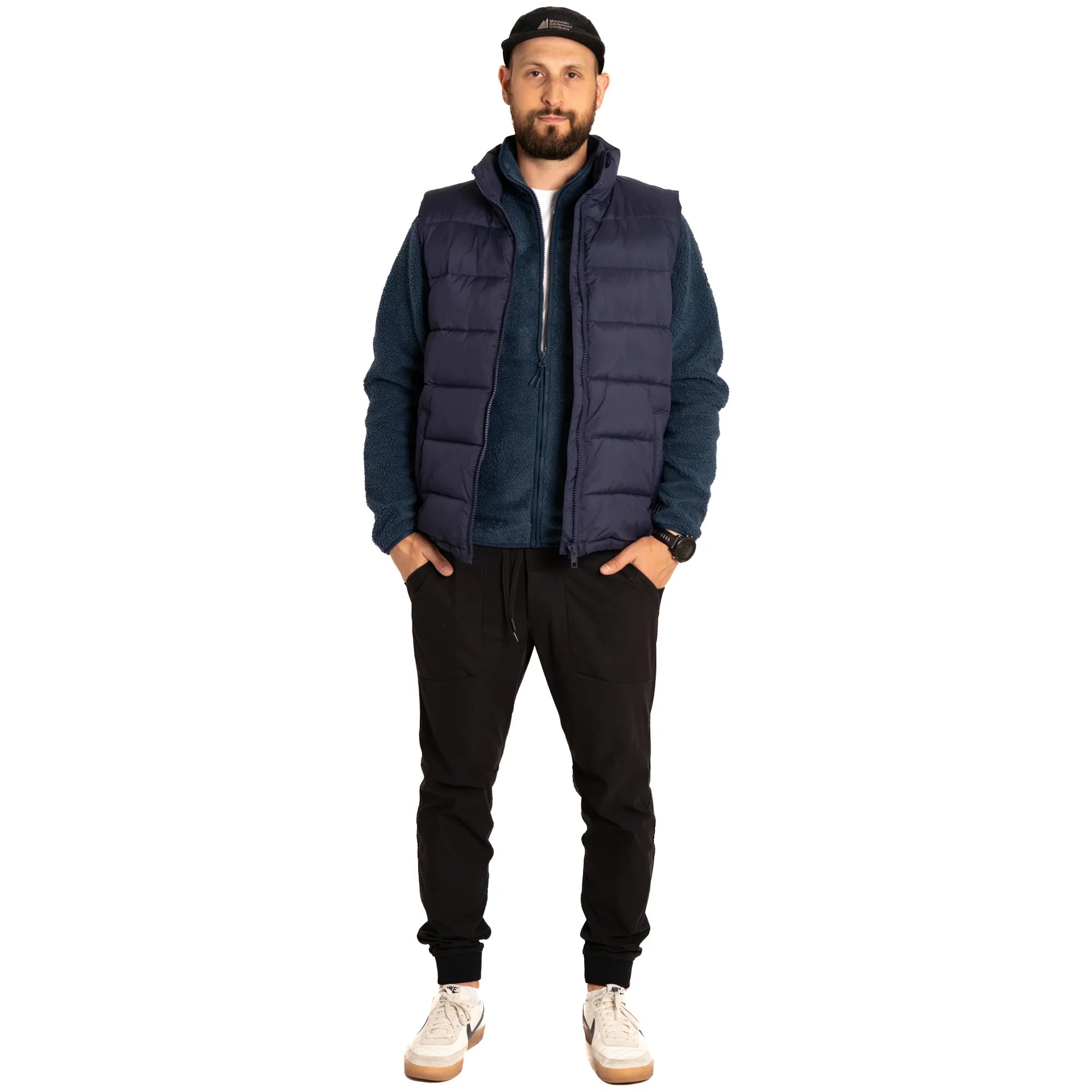 Man wearing navy puffer vest, blue zip-up jacket, black joggers, white sneakers, and cap