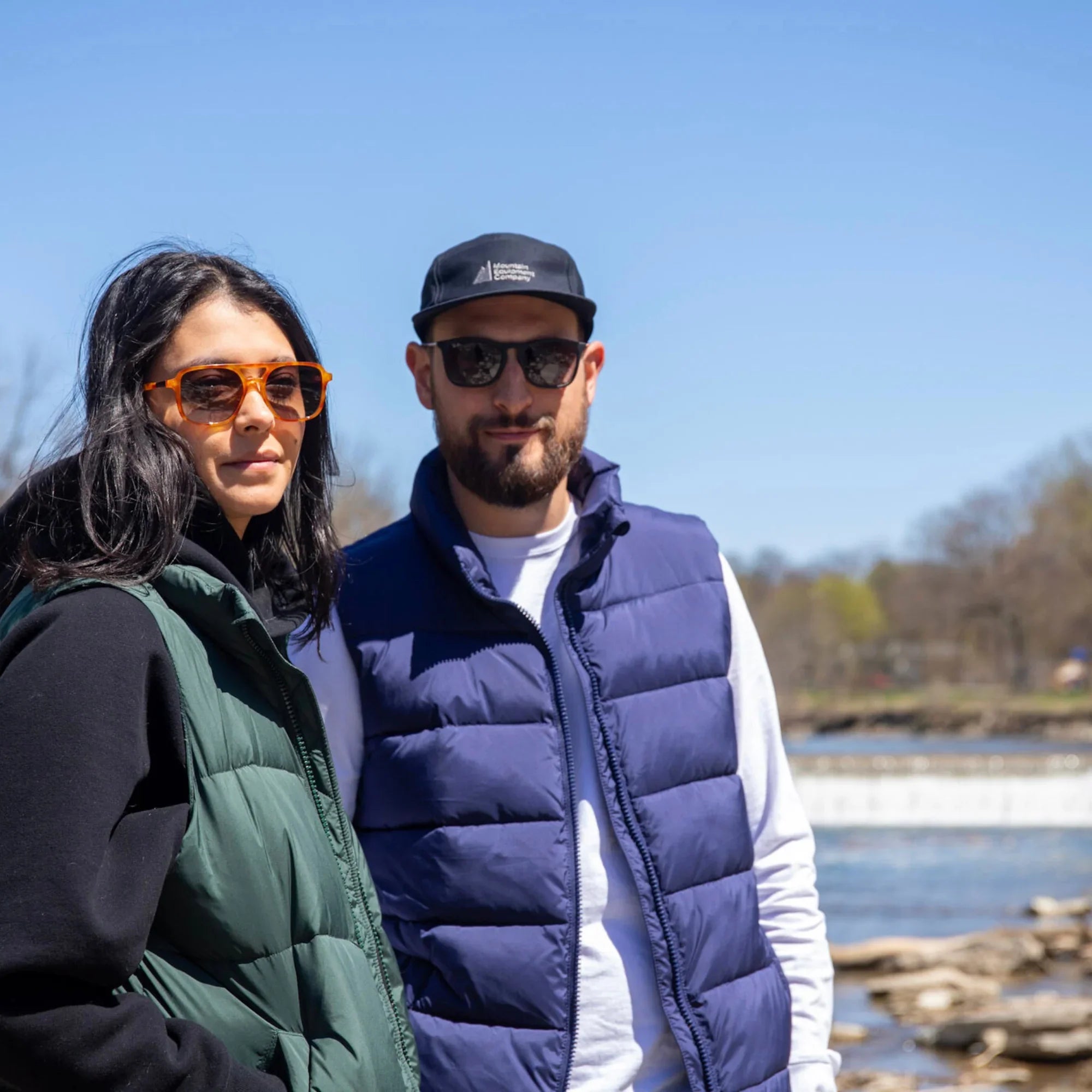 Two people wearing puffer vests and sunglasses outdoors by a river on a sunny day
