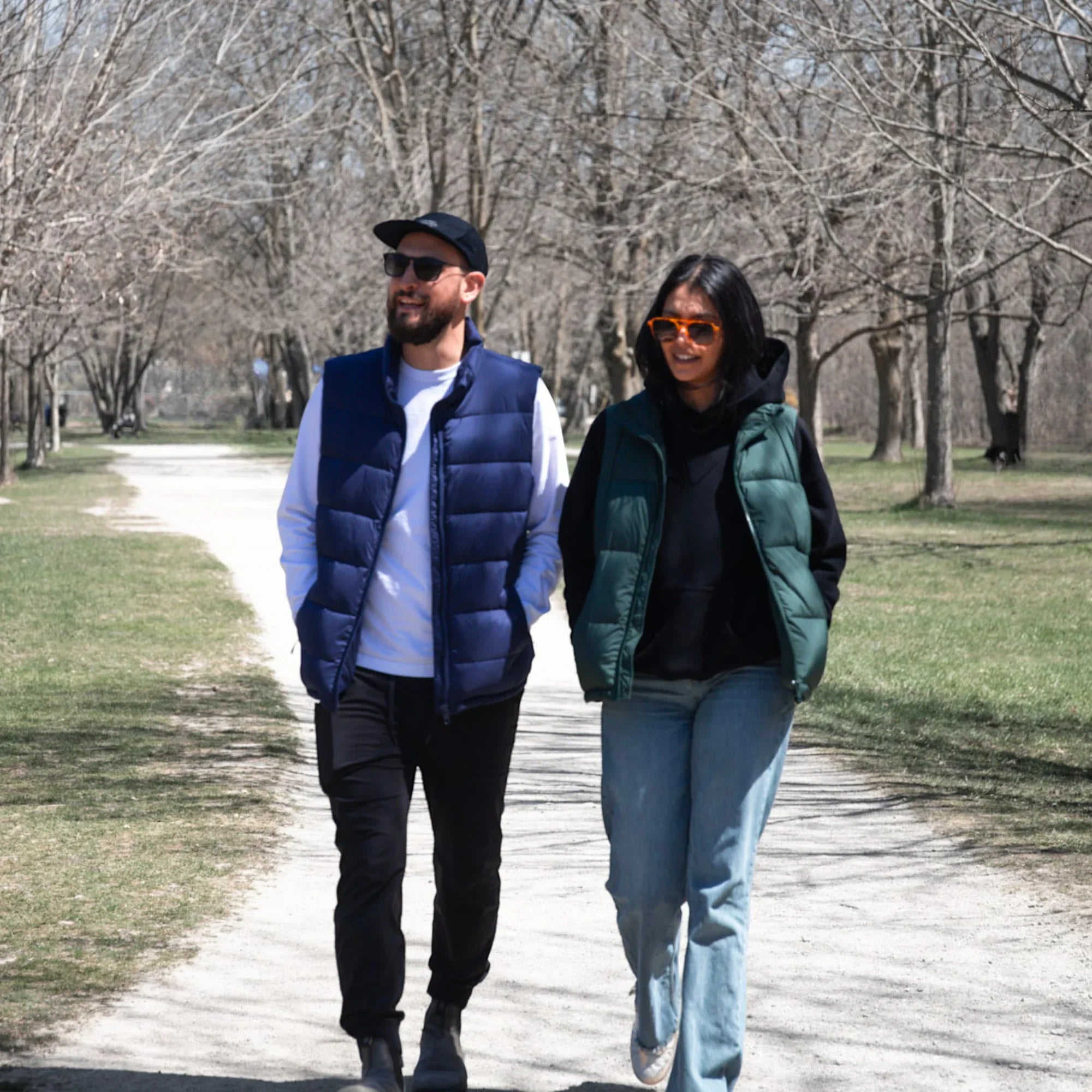 Couple wearing puffer vests and sunglasses walking on a park path in early spring