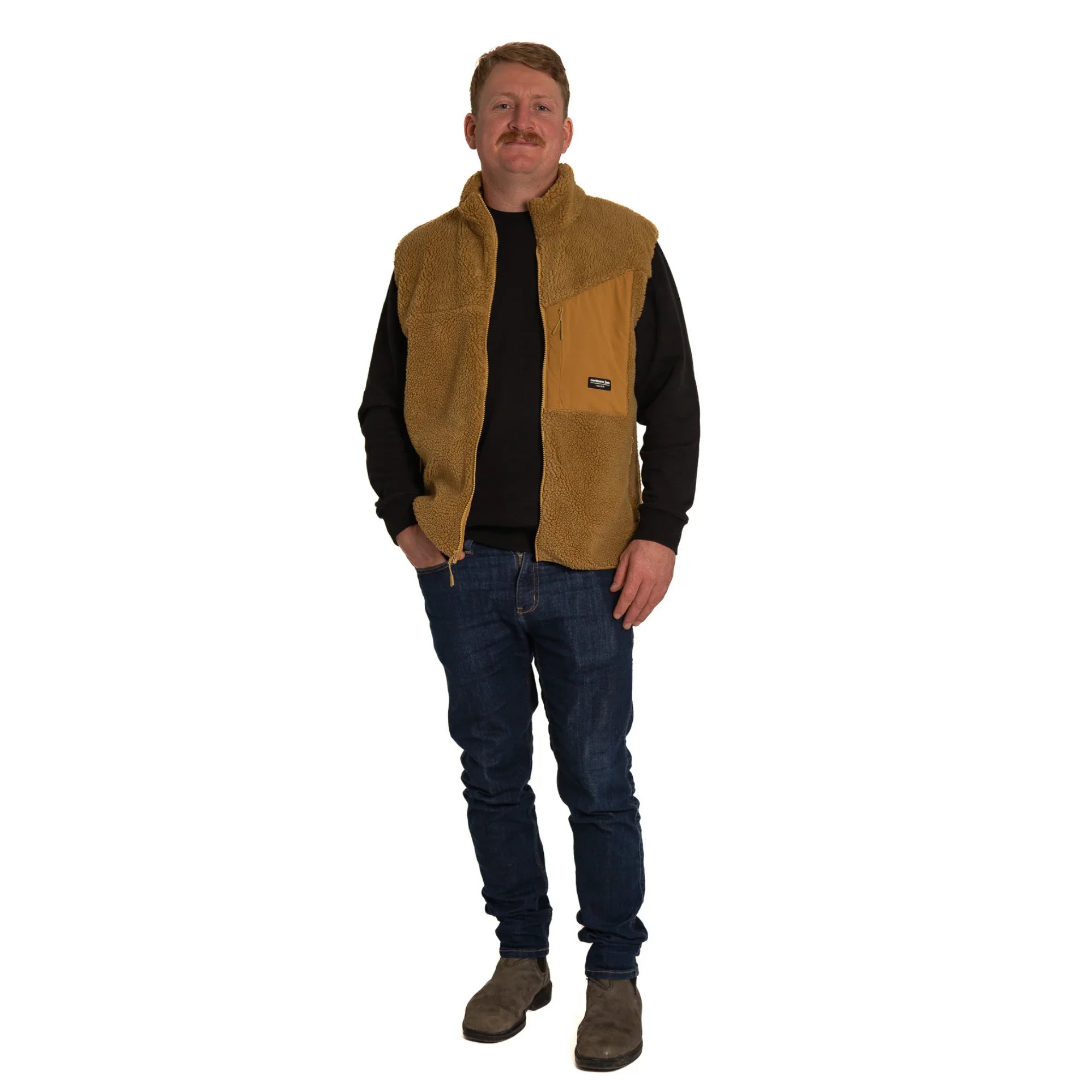 Man wearing tan fleece vest, black long sleeve shirt, jeans, and boots on white background
